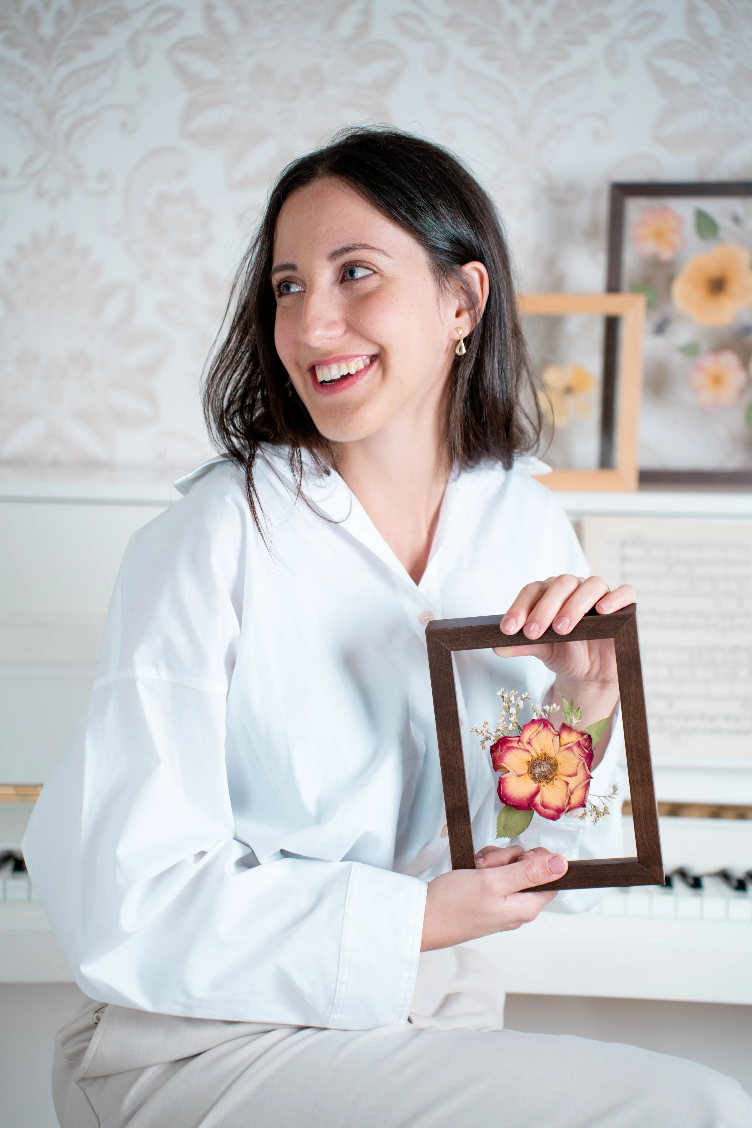 A woman with dark hair, wearing a white shirt, smiling and sitting while holding a wooden frame with a flower arrangement inside. The background features floral patterned wallpaper and framed artwork.