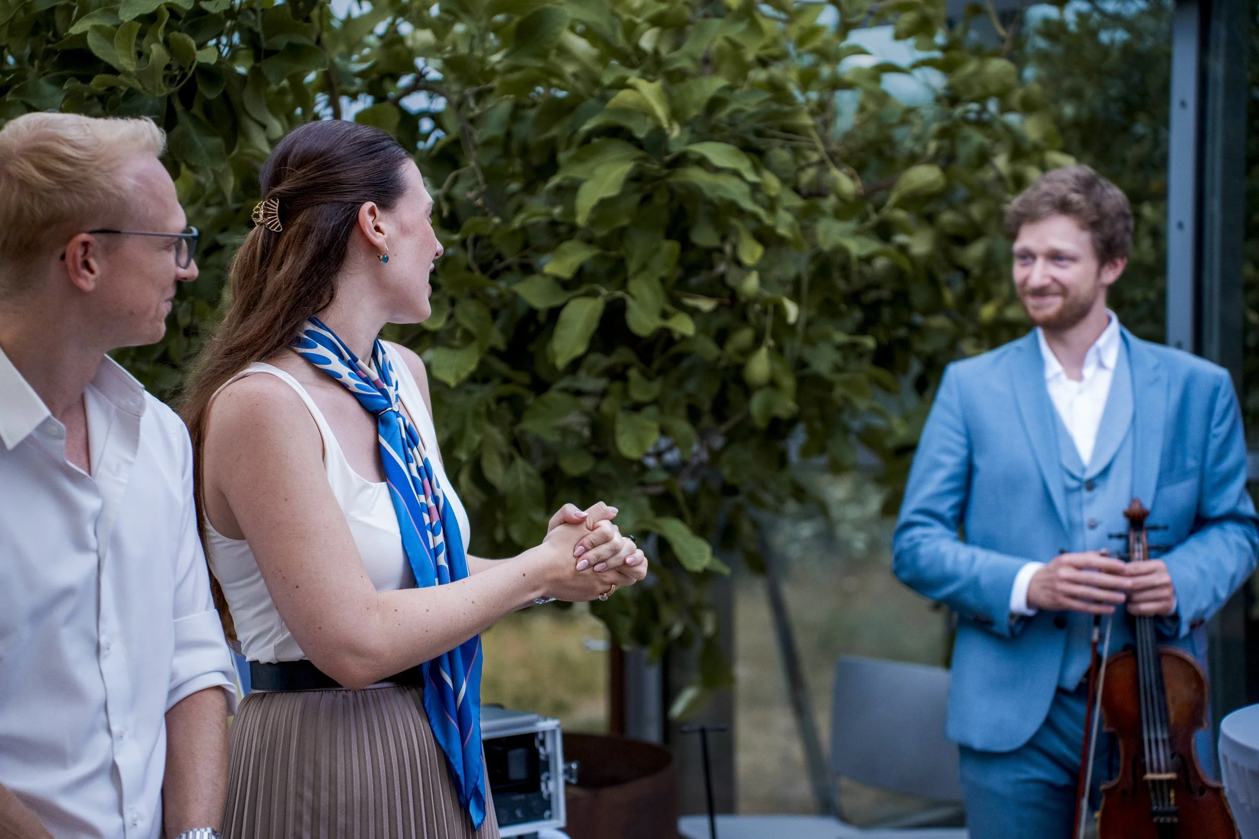 Four people at an outdoor event: two men and one woman, one woman is shaking hands with a young man in a blue suit with a violin. The background features green trees.