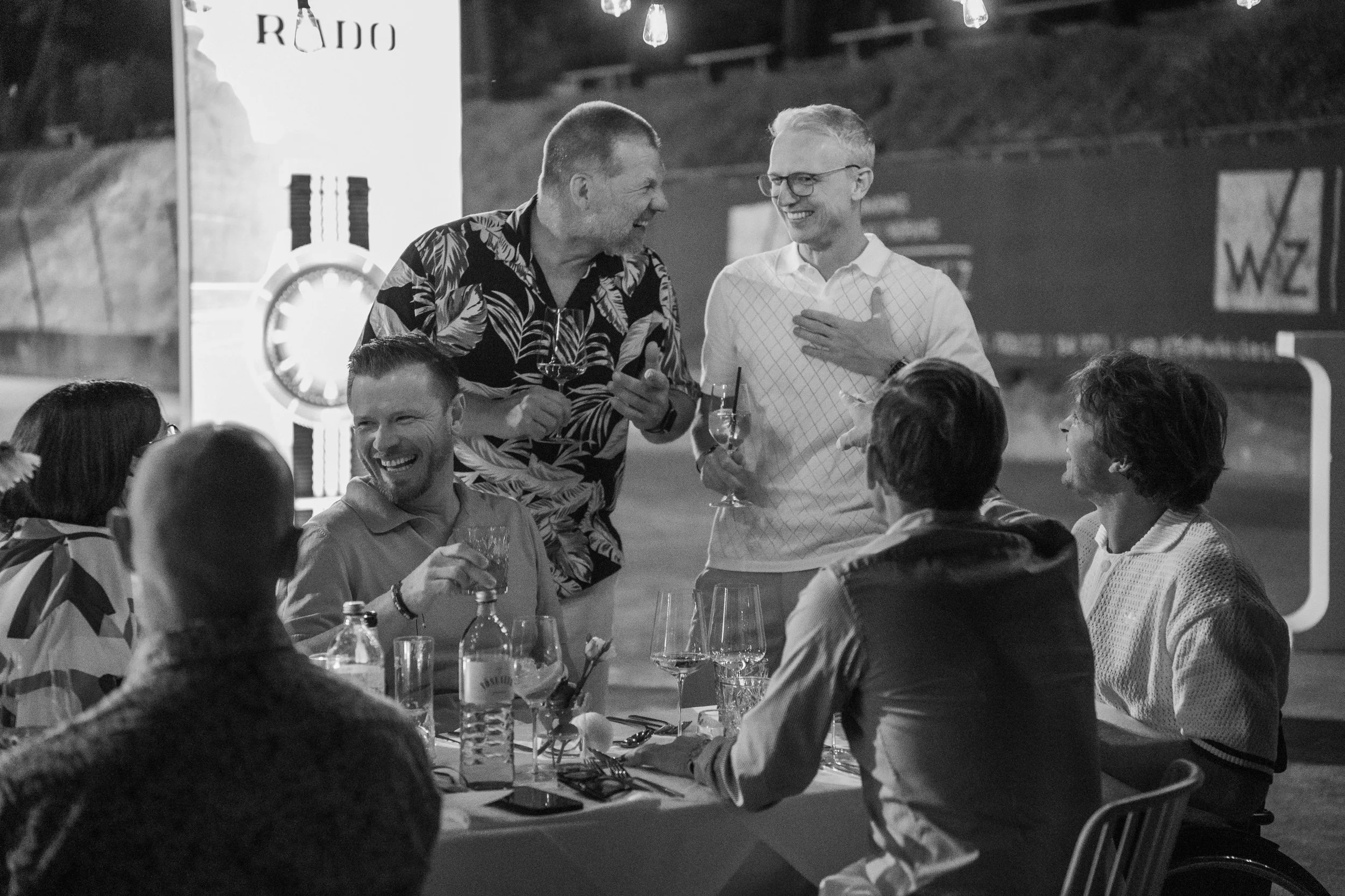People dining at a restaurant, with two men standing and talking, one holding a glass, and others seated around a table with drinks and bottles, enjoying conversation.