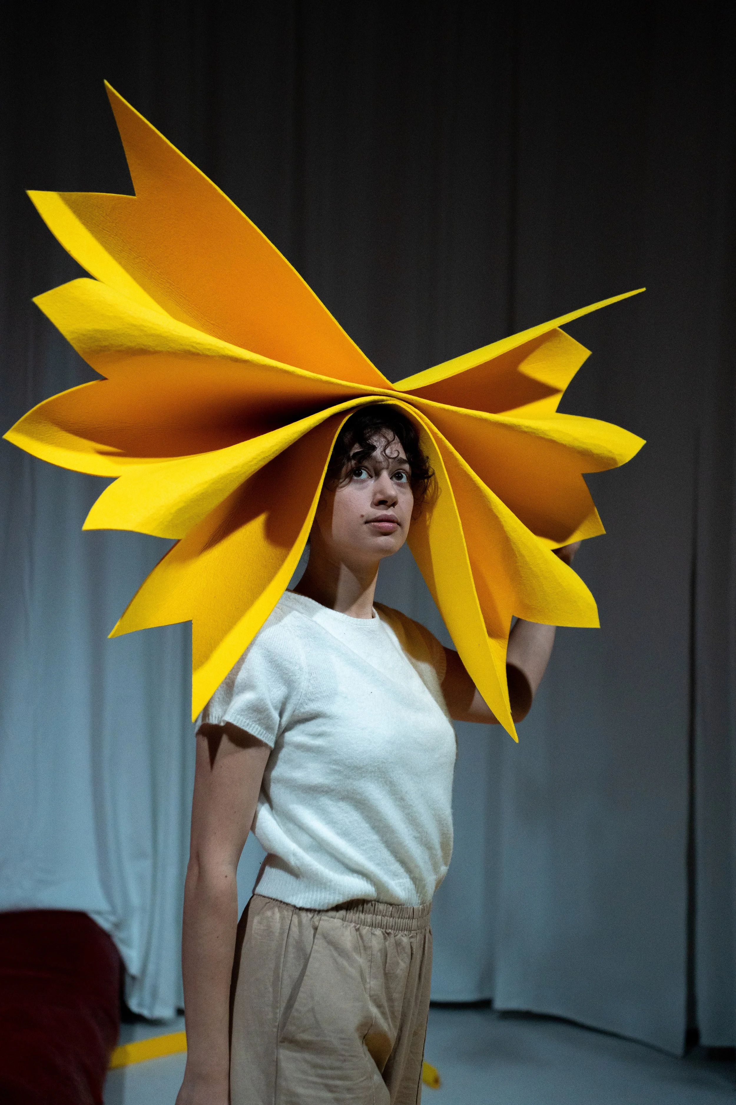 A person wears a large yellow flower-shaped costume around their head, standing indoors against a gray wall, with only their upper body visible.