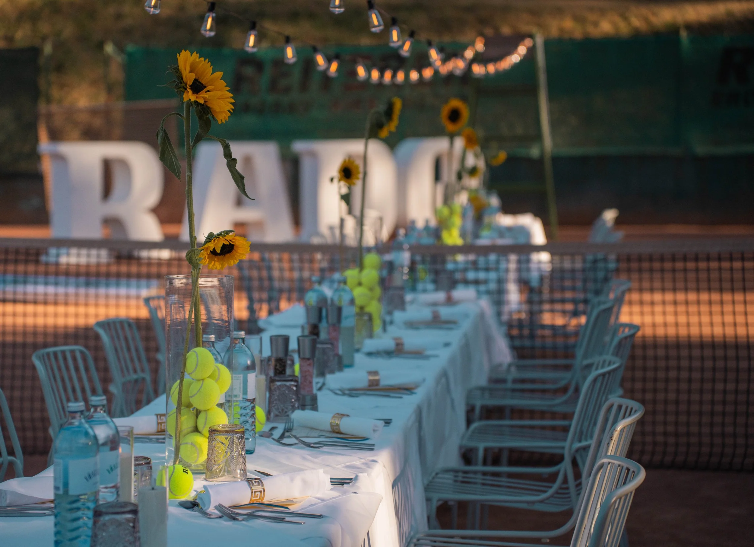 A long outdoor table set for a meal with white tablecloths, water bottles, tennis balls, and sunflowers in glass vases, with chairs and tennis balls hanging from the flowers, at dusk or evening.