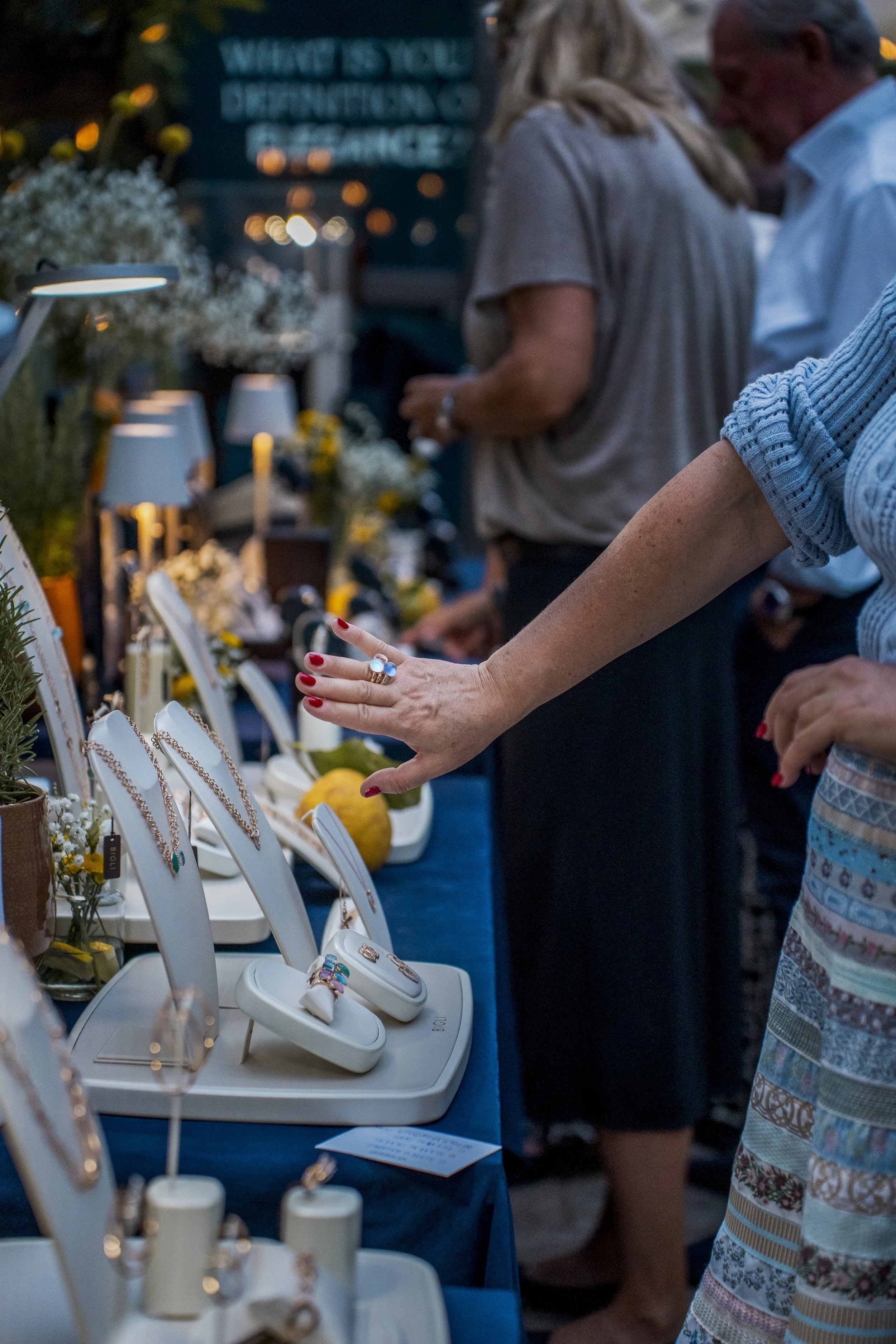People browsing jewelry at an outdoor market stall display with necklaces and rings on white stands.
