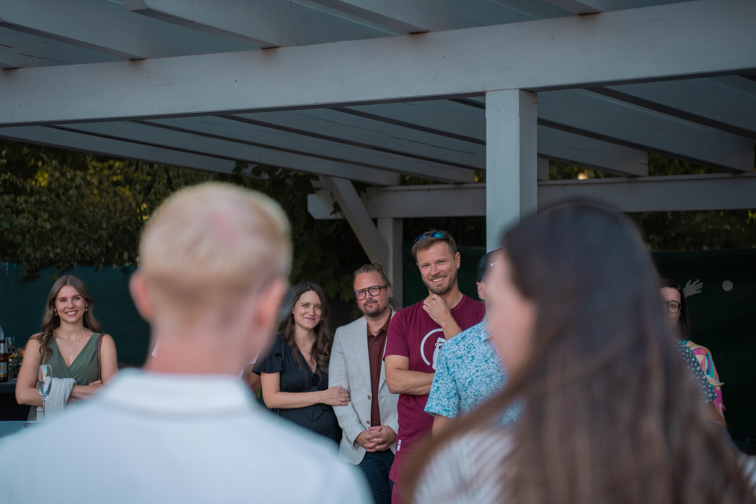 Group of people standing outdoors under a structure with a metal roof, engaged in conversation at a social gathering.