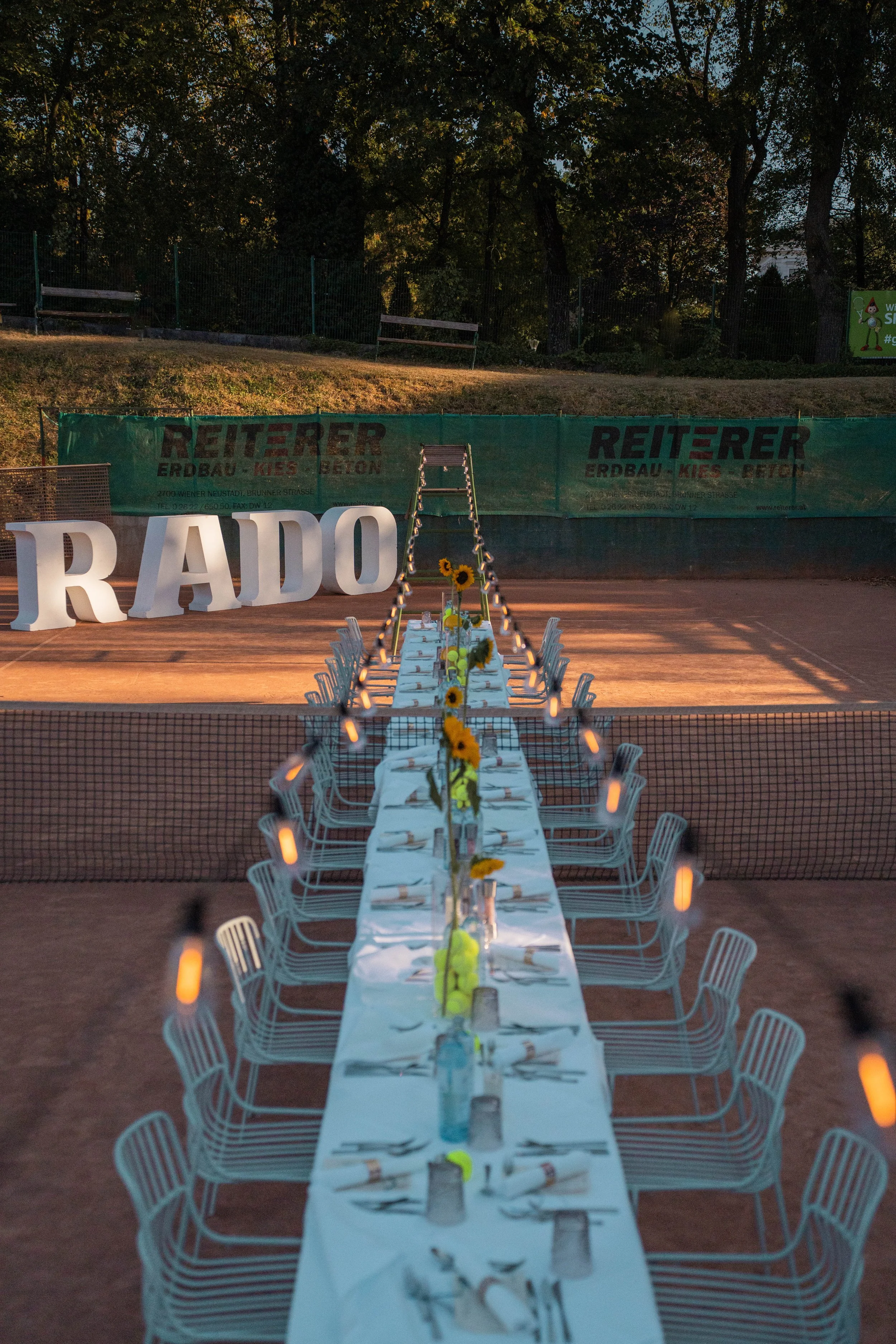Long outdoor dining table set for a meal, with chairs on either side, decorated with small vases of sunflowers and candles, located on a tennis court in the evening. In the background, large letters spell out 'RADO', and there is a green fence with advertisements and trees behind it.