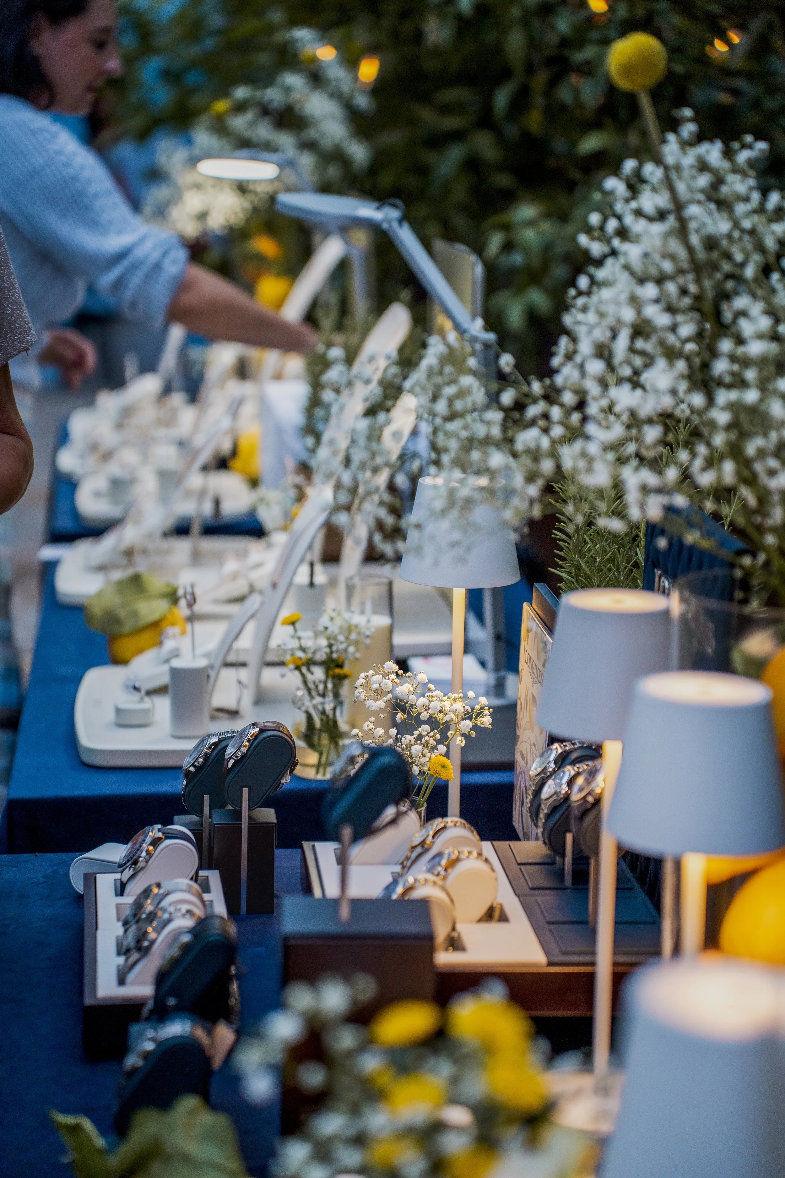 A jewelry store display with various watches, jewelry, and decorative flowers, illuminated by table lamps and ambient lighting.