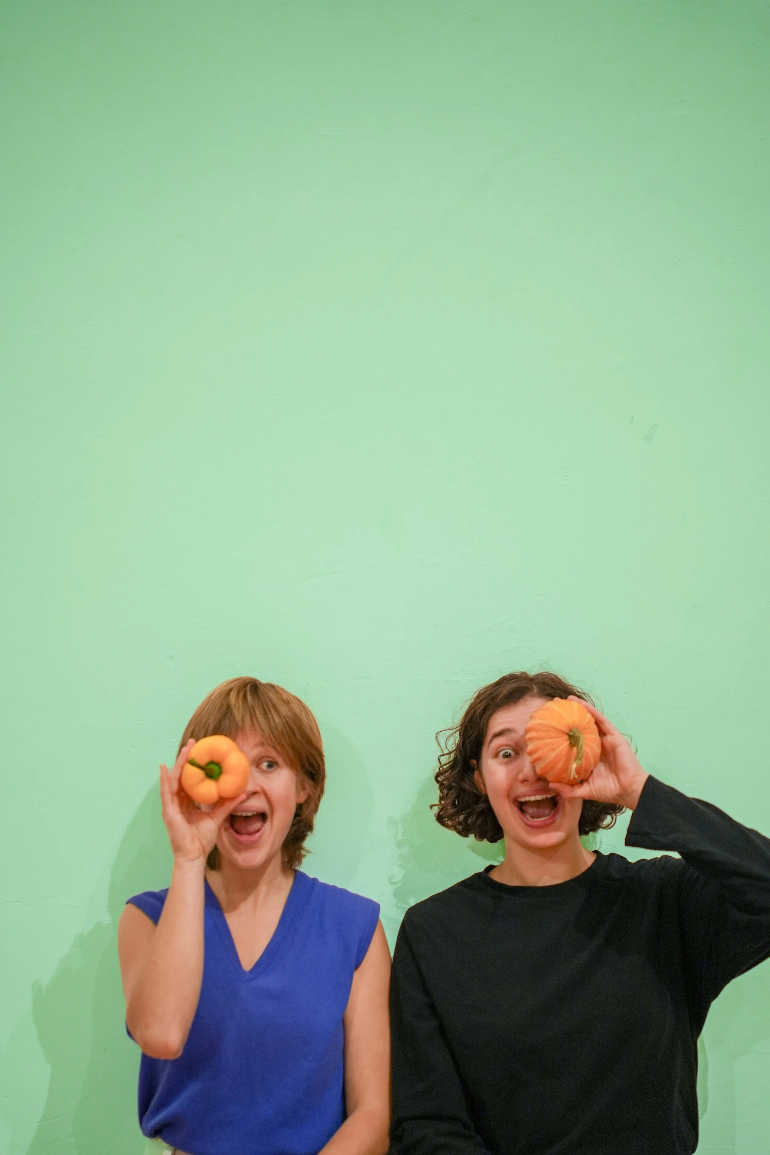 Two women are holding small pumpkins up to their eyes and smiling. They stand against a plain green background.