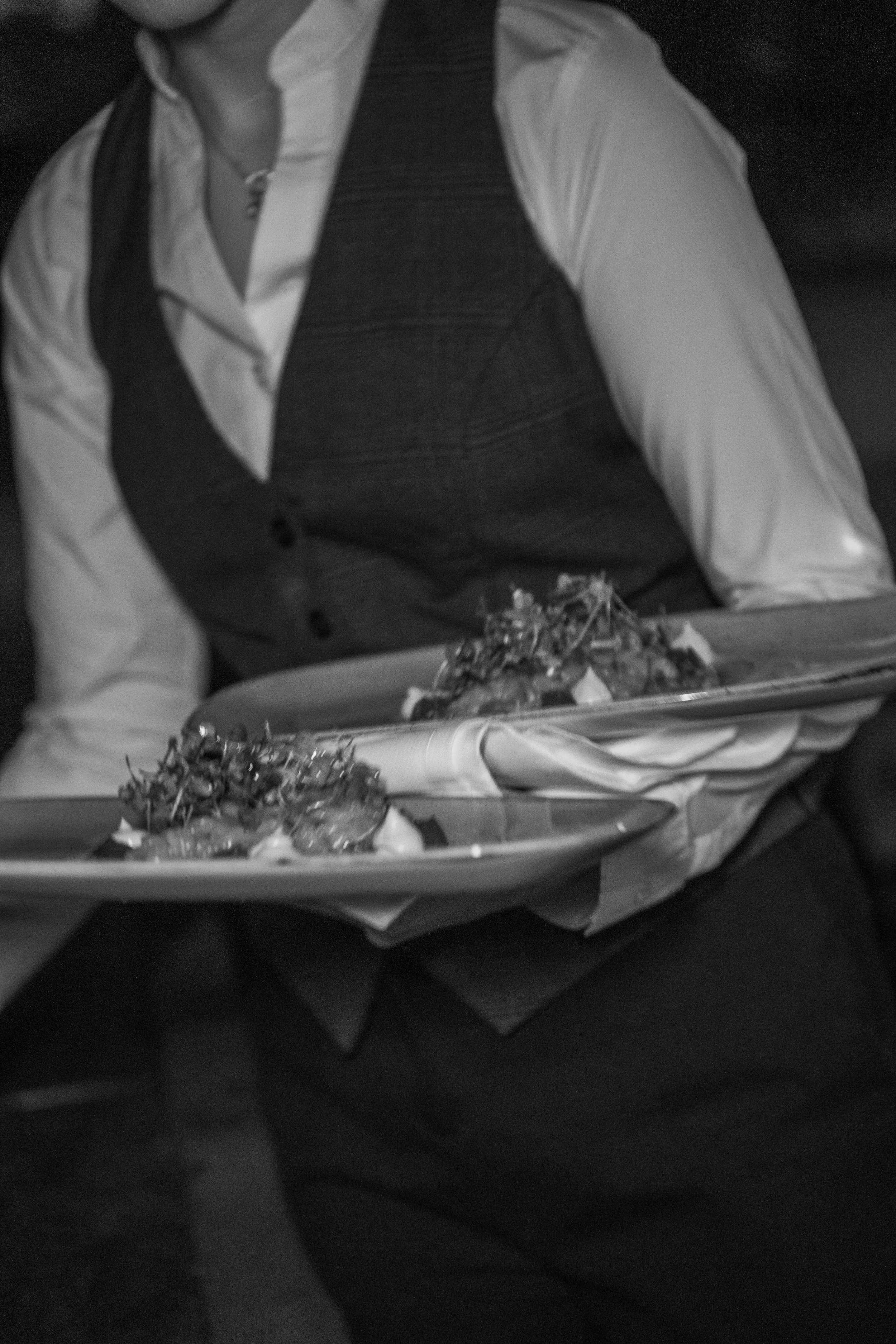 Person wearing a white shirt and dark vest holding a tray with food, possibly at a celebration or event.