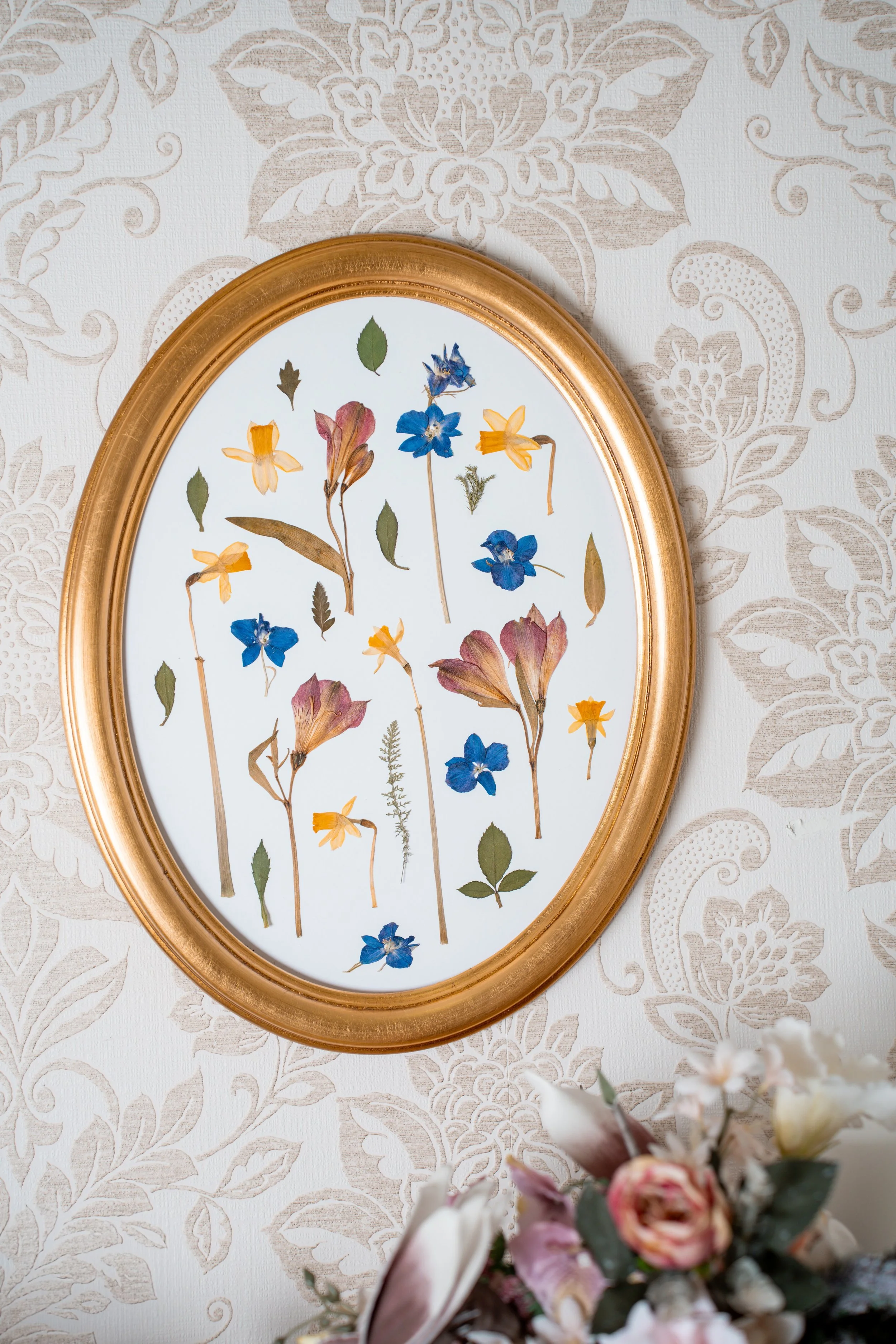 An oval framed botanical art piece with pressed flowers and leaves on a white background, mounted on a textured, patterned wall.