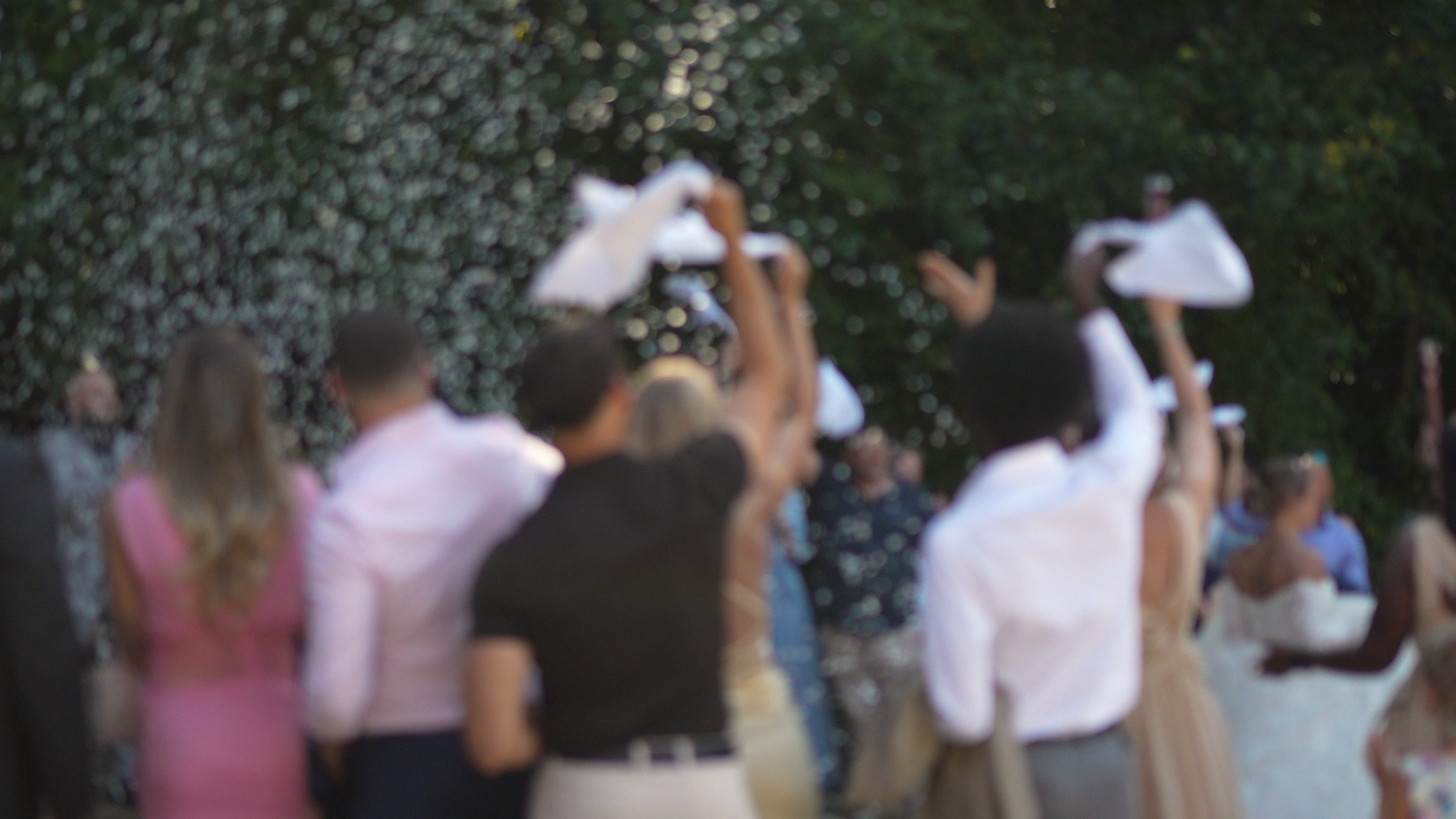 A large group of people outdoors during daytime, some holding white napkins in celebration, gathered in front of trees.