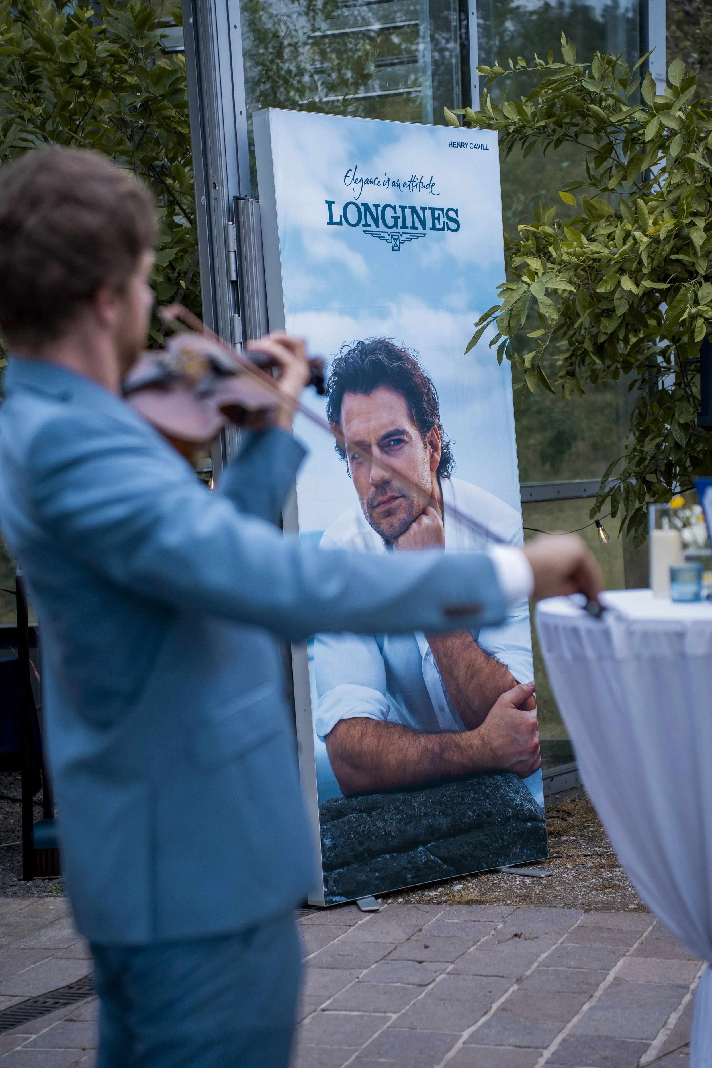 A man playing a violin with a large advertisement for Longines watches in the background, featuring a model with a serious expression, sitting with his hand on his chin.