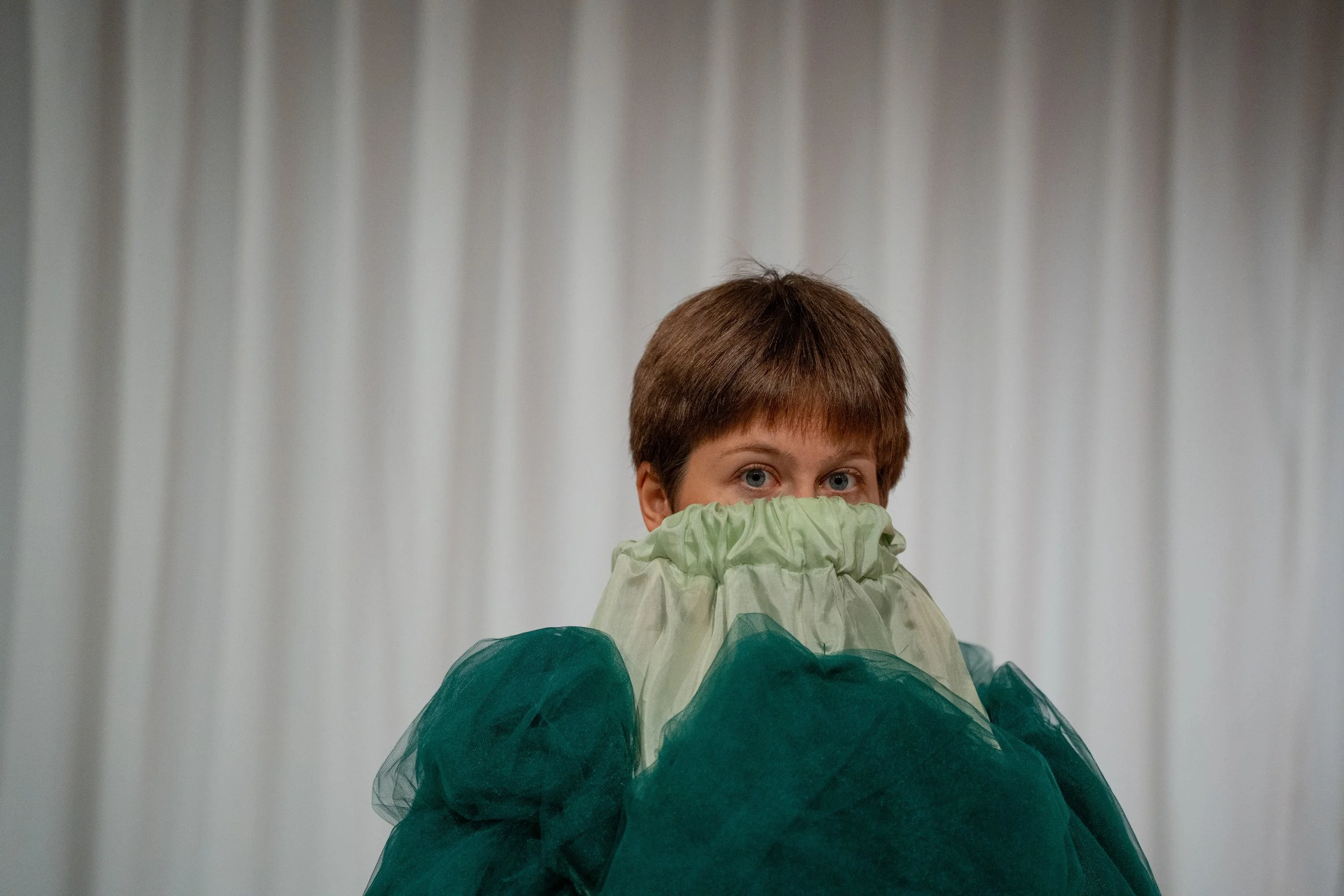 A person with short brown hair and blue eyes looks over their shoulder, wearing a light green jacket with a dark green bottom layer and a green tulle skirt. The background is a plain, light-colored curtain.