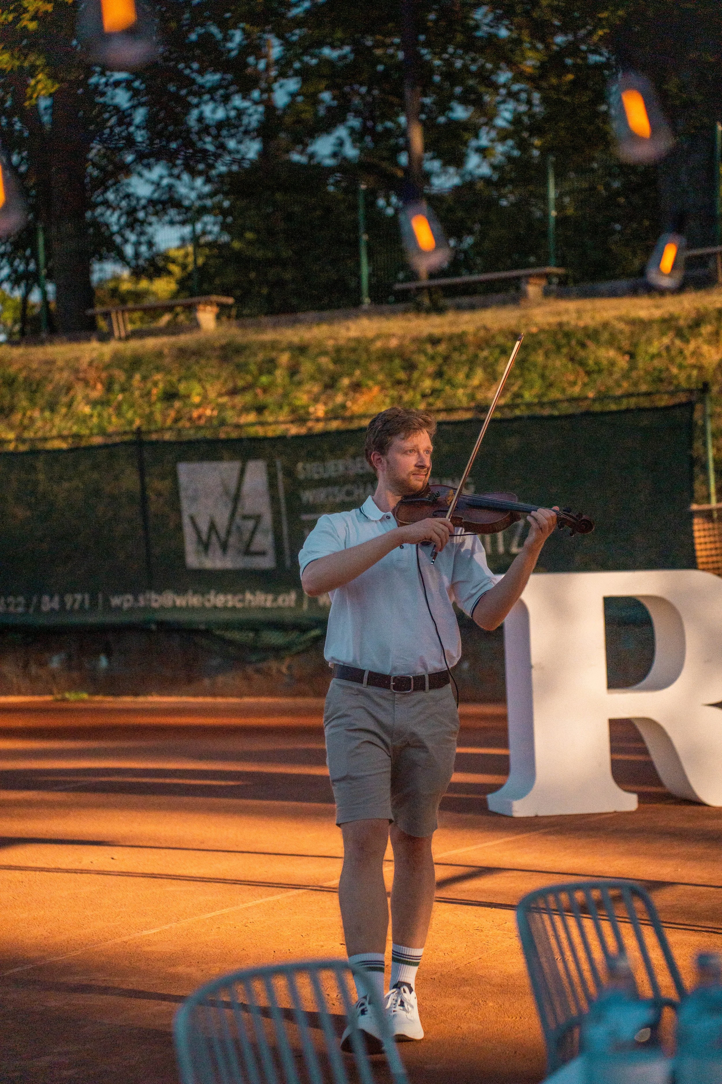 A man in a white shirt, beige shorts, white sneakers, and striped socks playing a violin on an outdoor stage at dusk or evening.