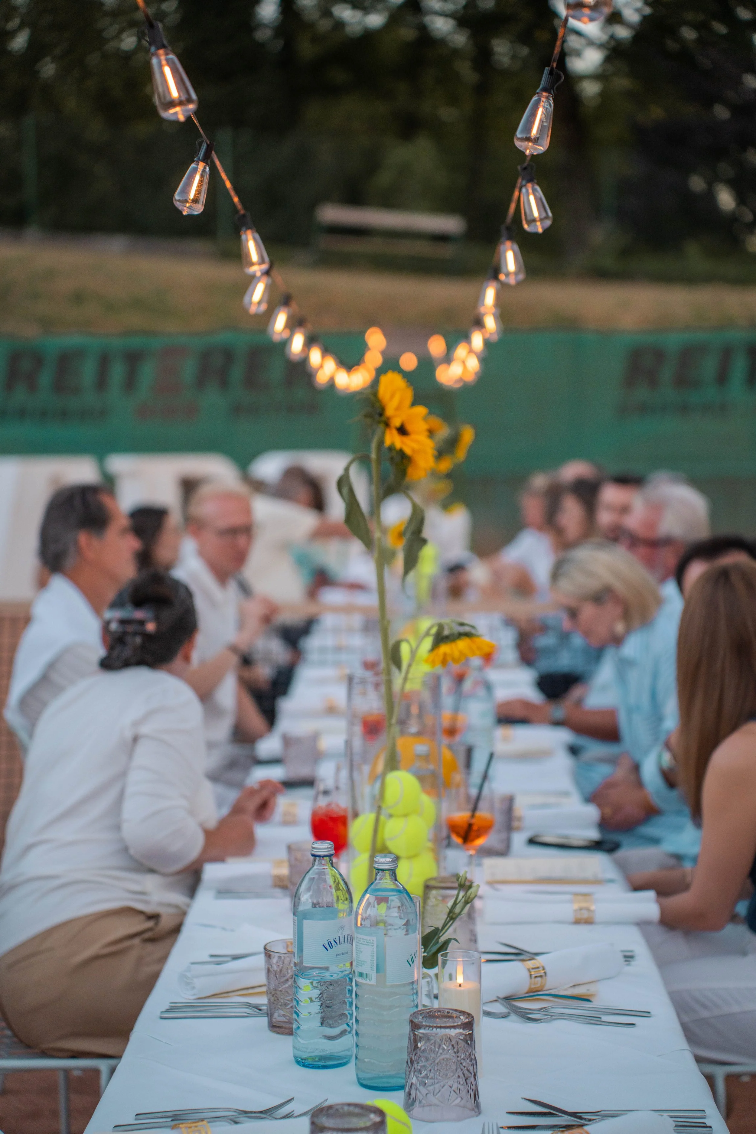People dining at a long outdoor table during evening, with a decorative string of lights overhead, flowers in the center of the table, and water bottles on the table.