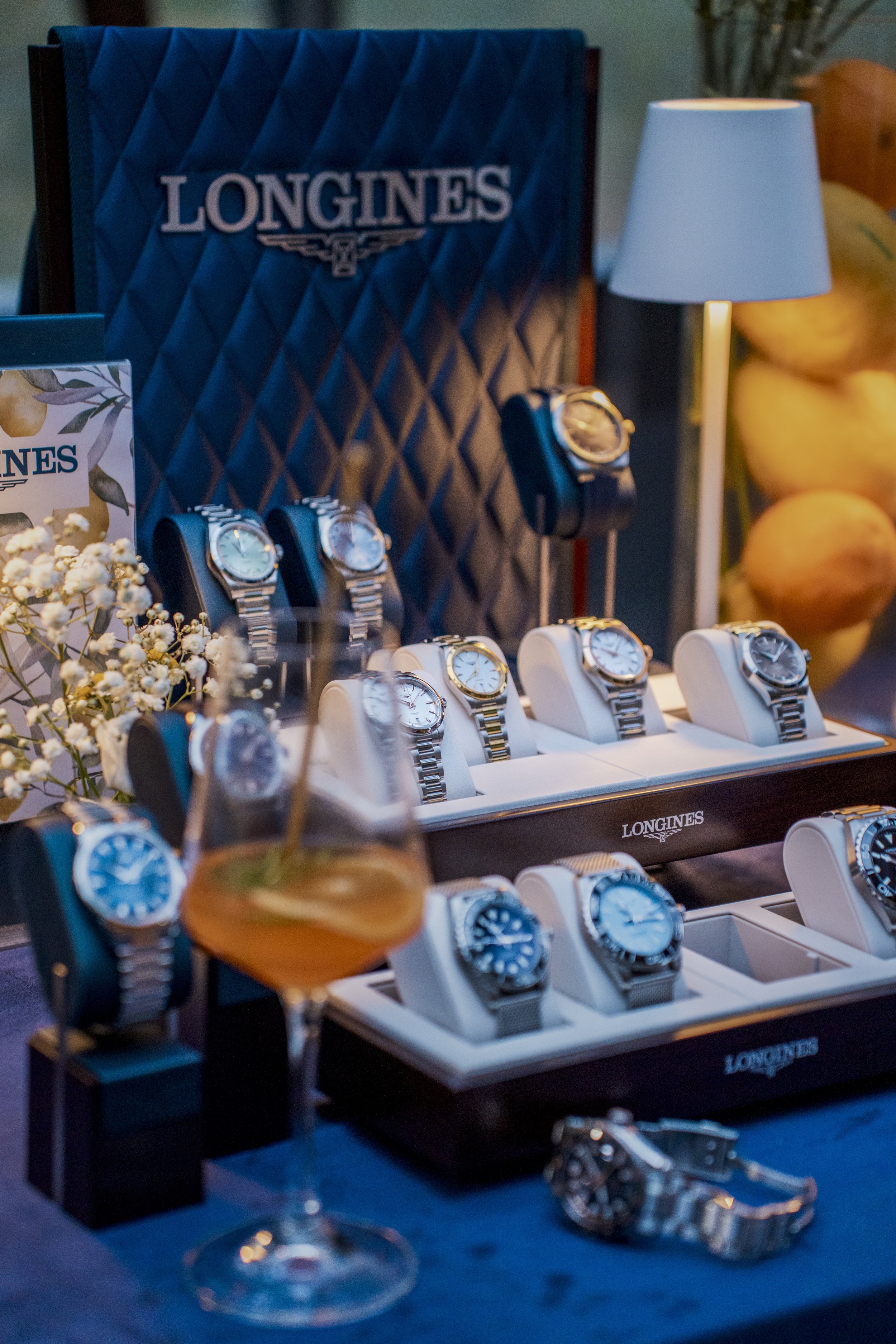 Display of various wristwatches on white stands and cushions, with a Longines branded display box and a table lamp in the background, and a drink in the foreground.