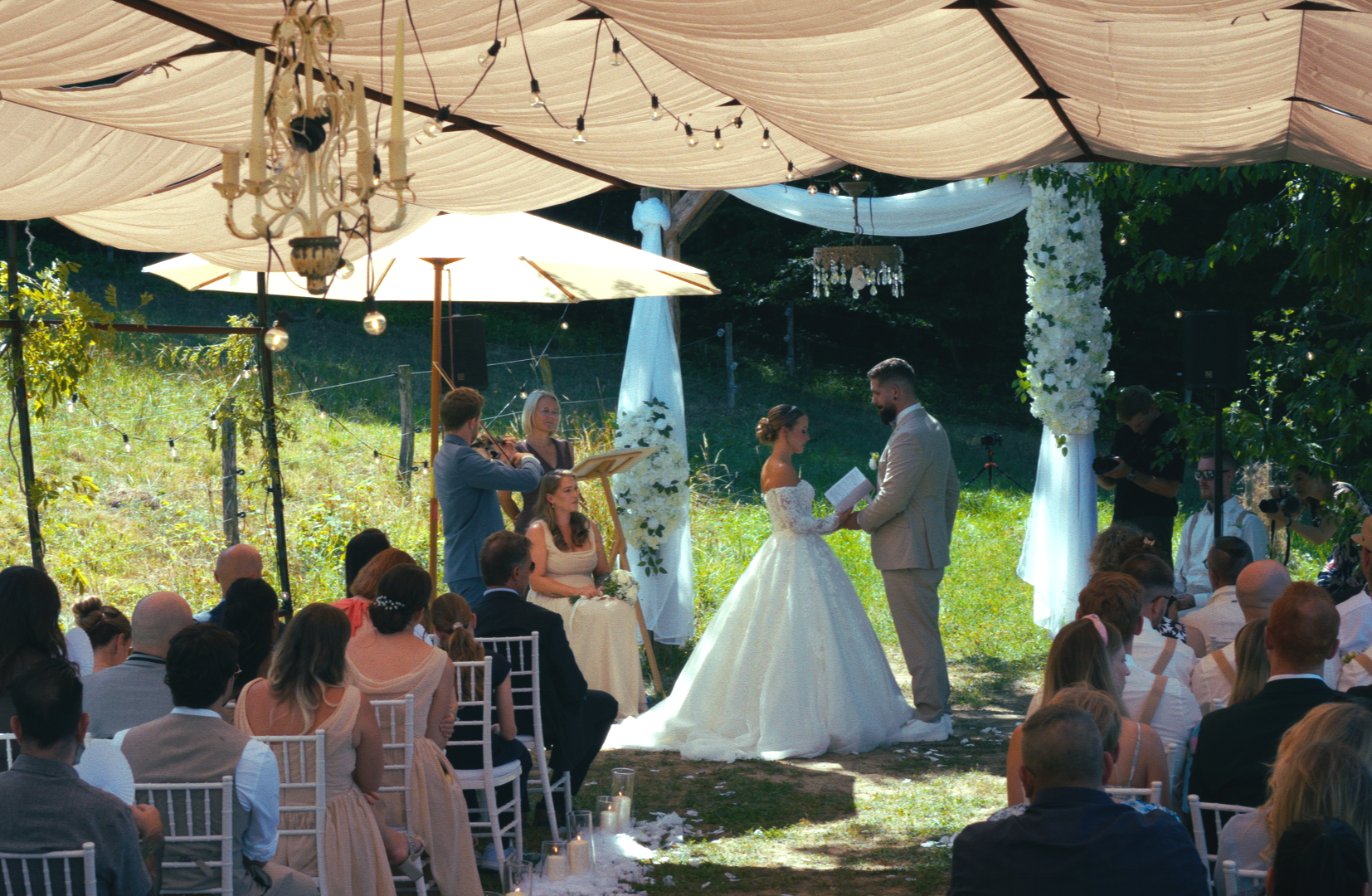 Wedding ceremony outdoors under a canopy of fabric and string lights. Bride and groom exchange vows, surrounded by seated guests, floral decorations, and musicians.