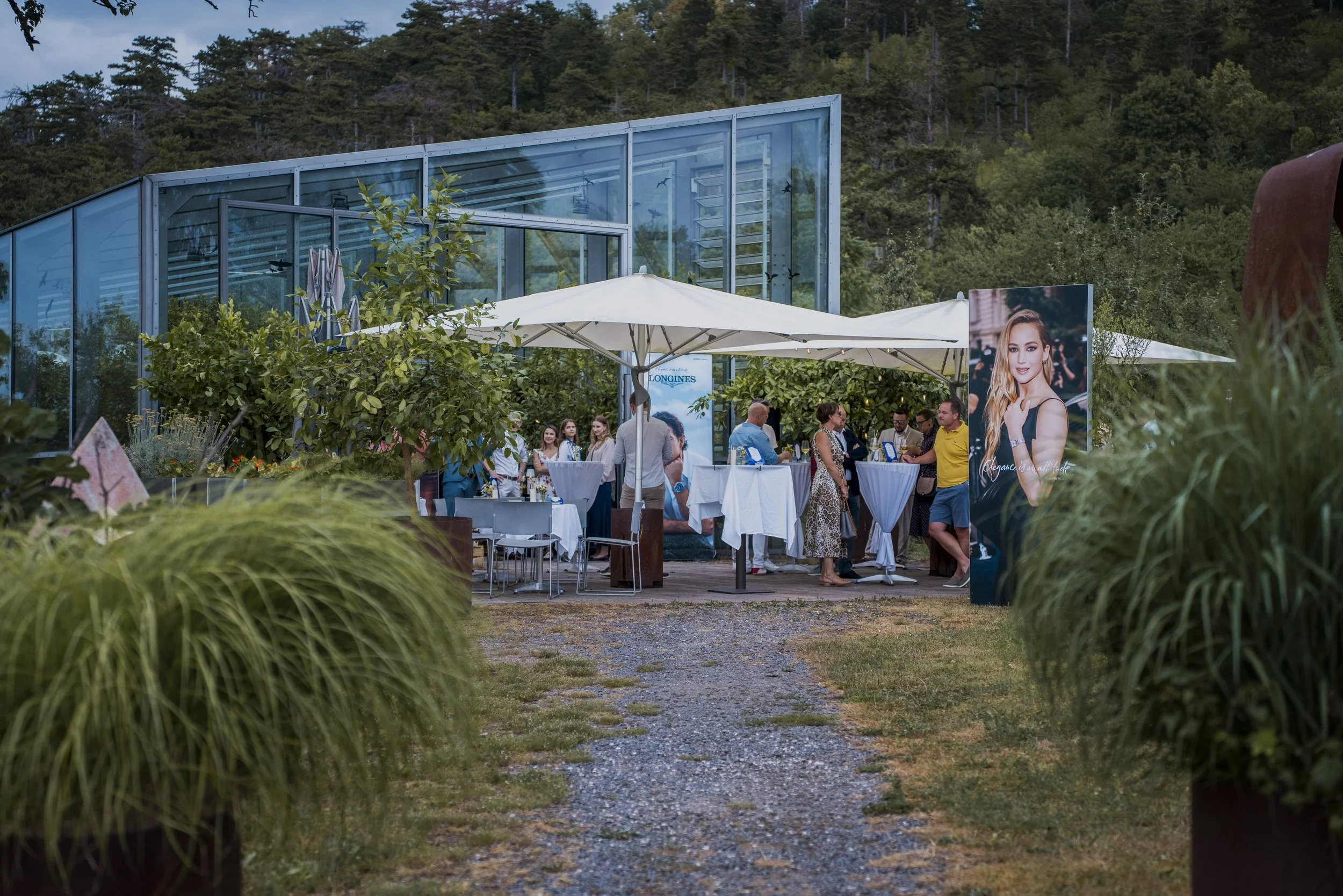 An outdoor reception area with white tables and umbrellas, surrounded by lush greenery and trees, adjacent to a modern glass building, with people socializing and a large advertisement featuring a woman.