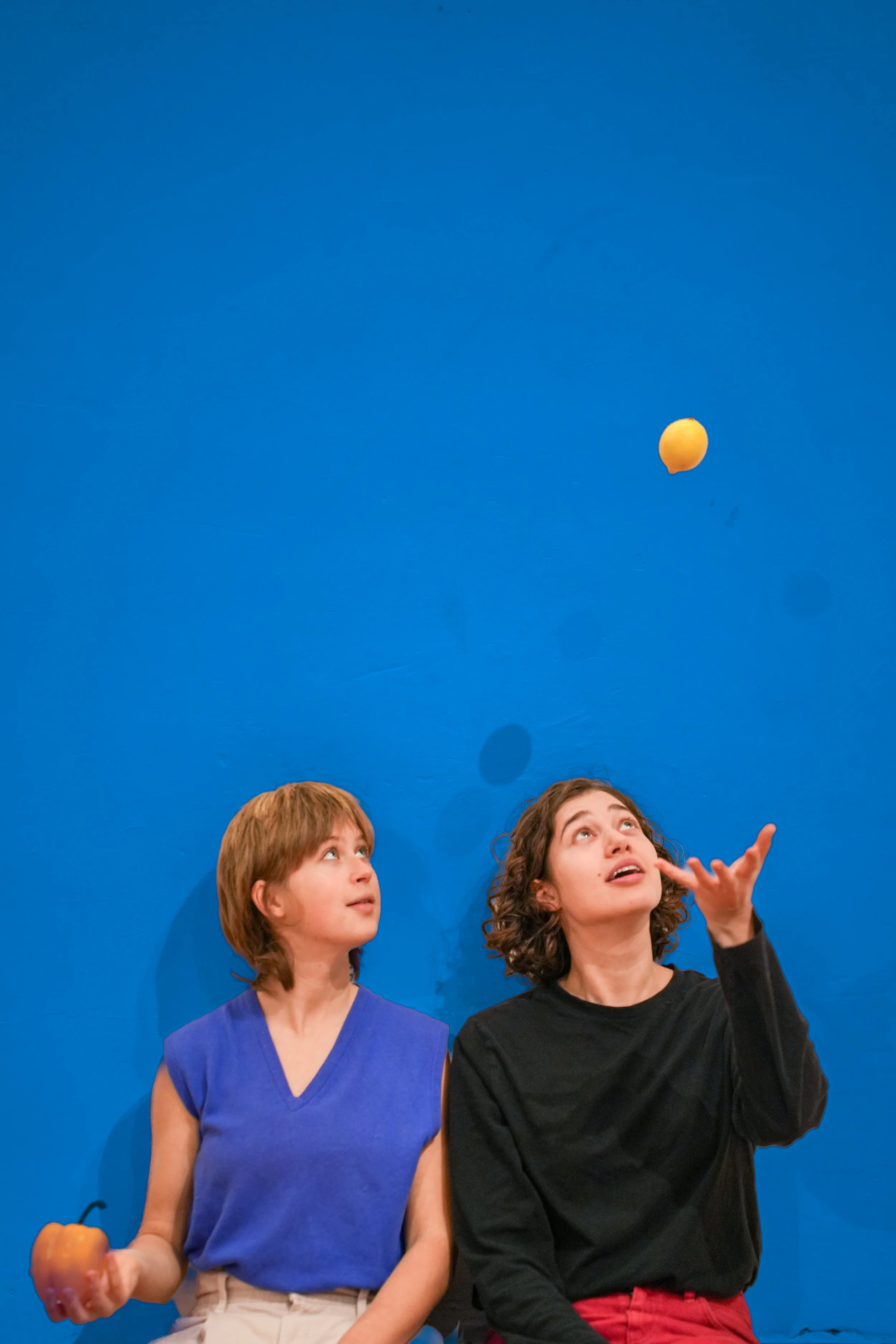 Two children with short brown hair and a woman with curly brown hair are sitting against a bright blue background. The woman is tossing a yellow ball into the air, and the boy with straight brown hair is holding an orange apple.