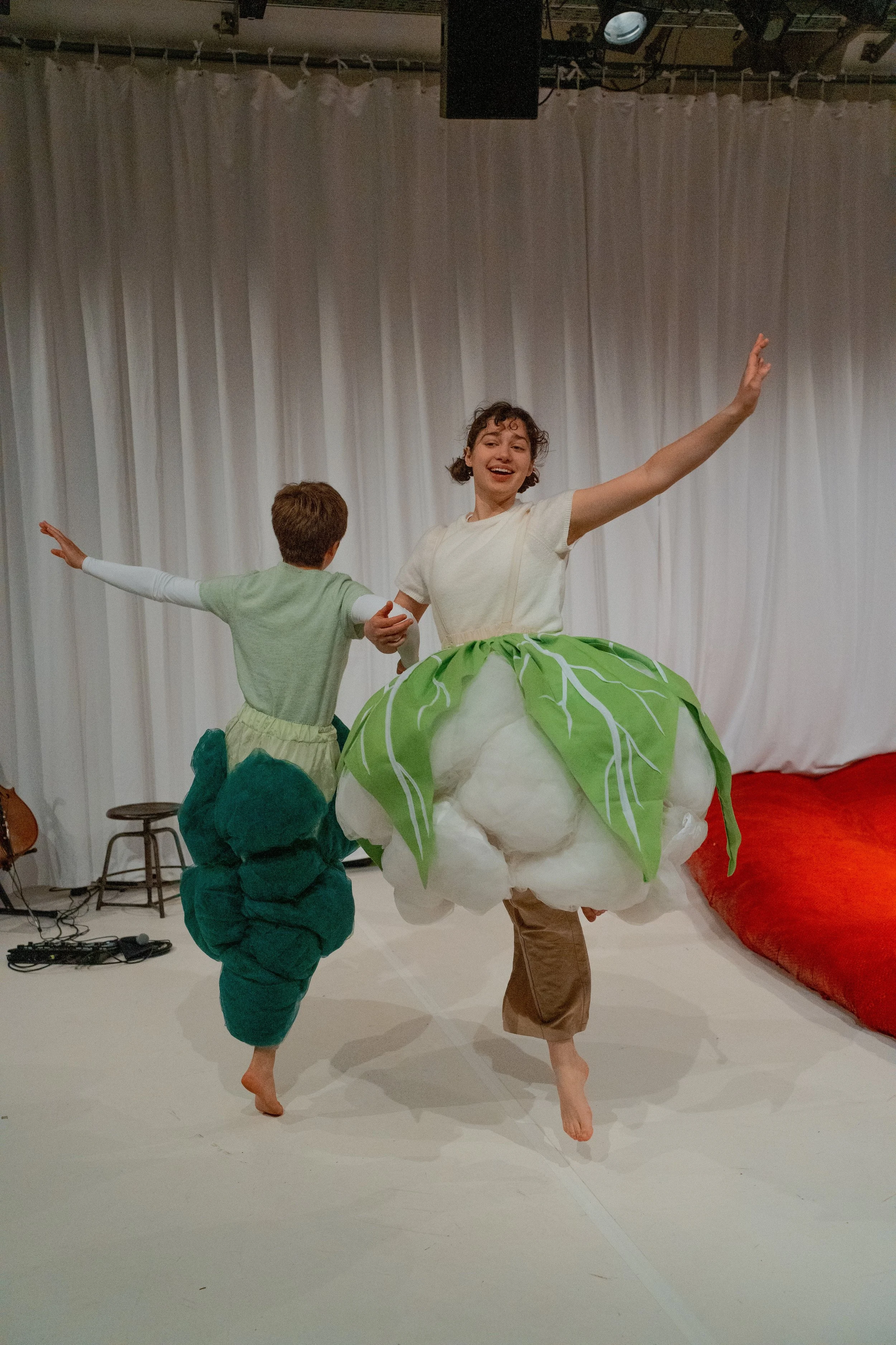 A person and a child performing a dance or theater act. The person is wearing a costume resembling a large, white flower with green leaves, while the child is dressed in a green and white outfit with a dinosaur or dragon head on it. They are barefoot