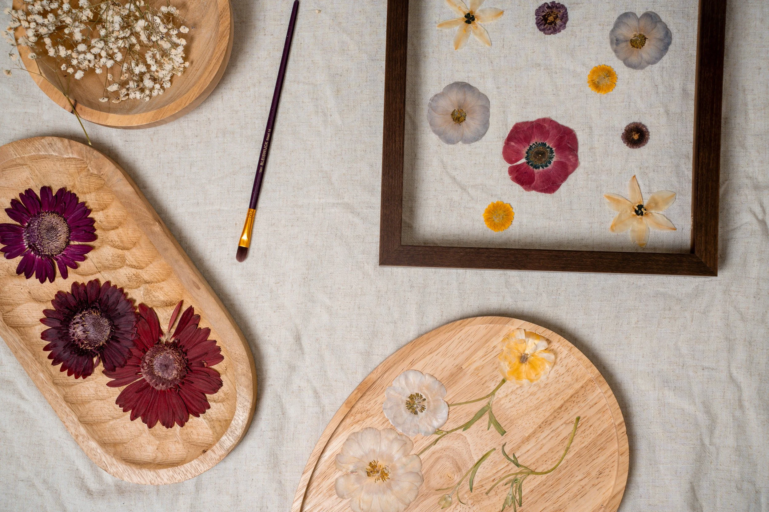 Flat lay of wooden plates and frames with pressed flowers on a light fabric background, a paintbrush with purple handle and yellow tip.