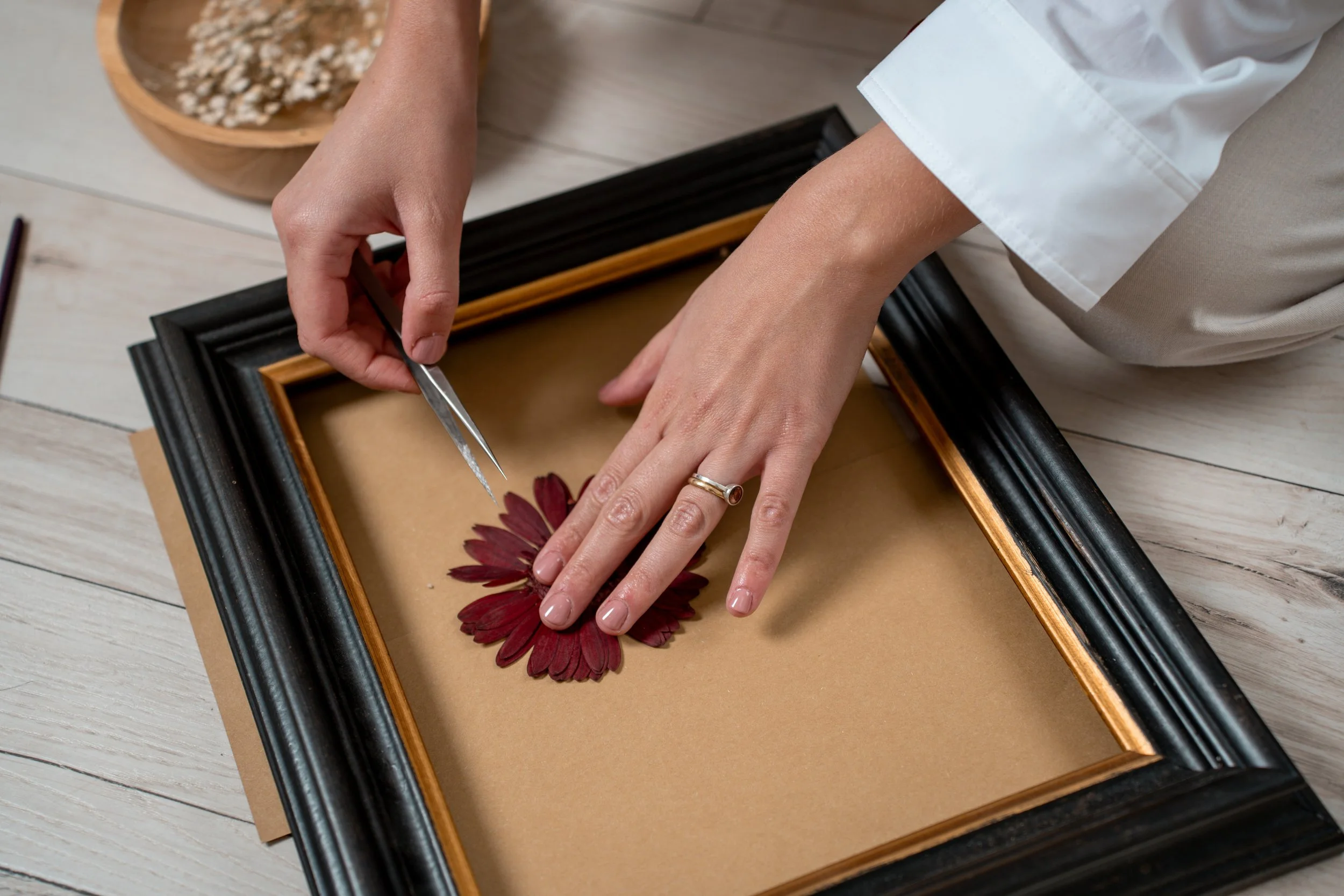 A person using tweezers to place or adjust a red flower in a shadow box frame. The shadow box has a black outer frame and a light brown inner background, and the person is wearing a white sleeve and a ring on their finger. There is a bowl with dried 