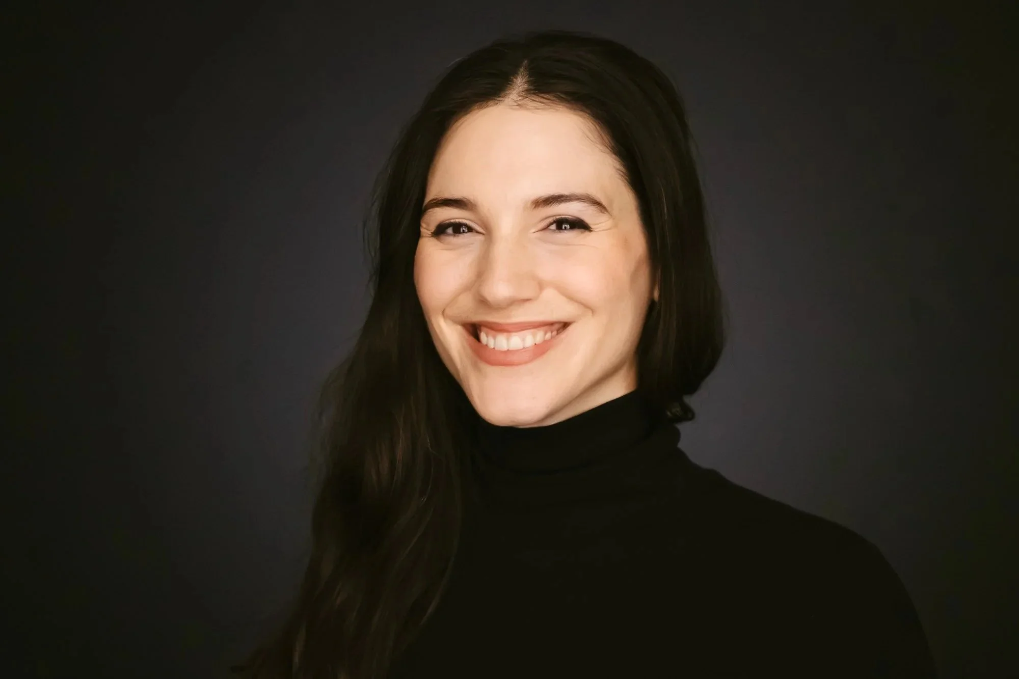 Danae Papadimitriou wearing a black turtleneck, is smiling and looking at the camera against a dark background.