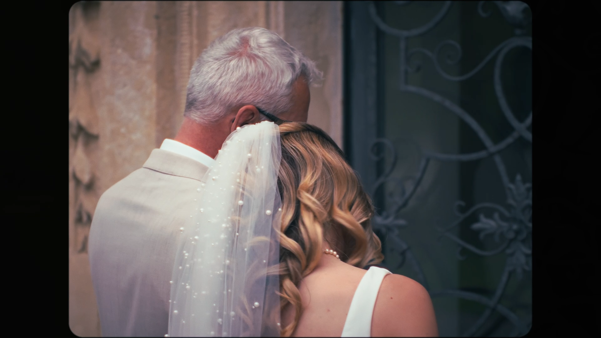 A bride and her father sharing a close moment, with the father's face pressed against the bride's head, and her hair styled in soft curls, wearing a wedding dress with pearl details, standing beside a decorative wrought-iron door.