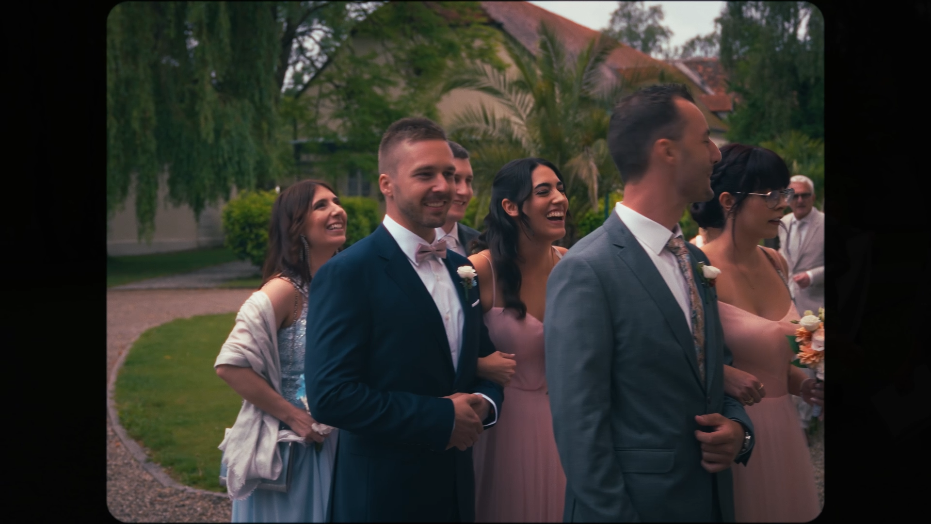 Group of people dressed in formal attire at an outdoor wedding or celebration, standing on a pathway with greenery and trees in the background.