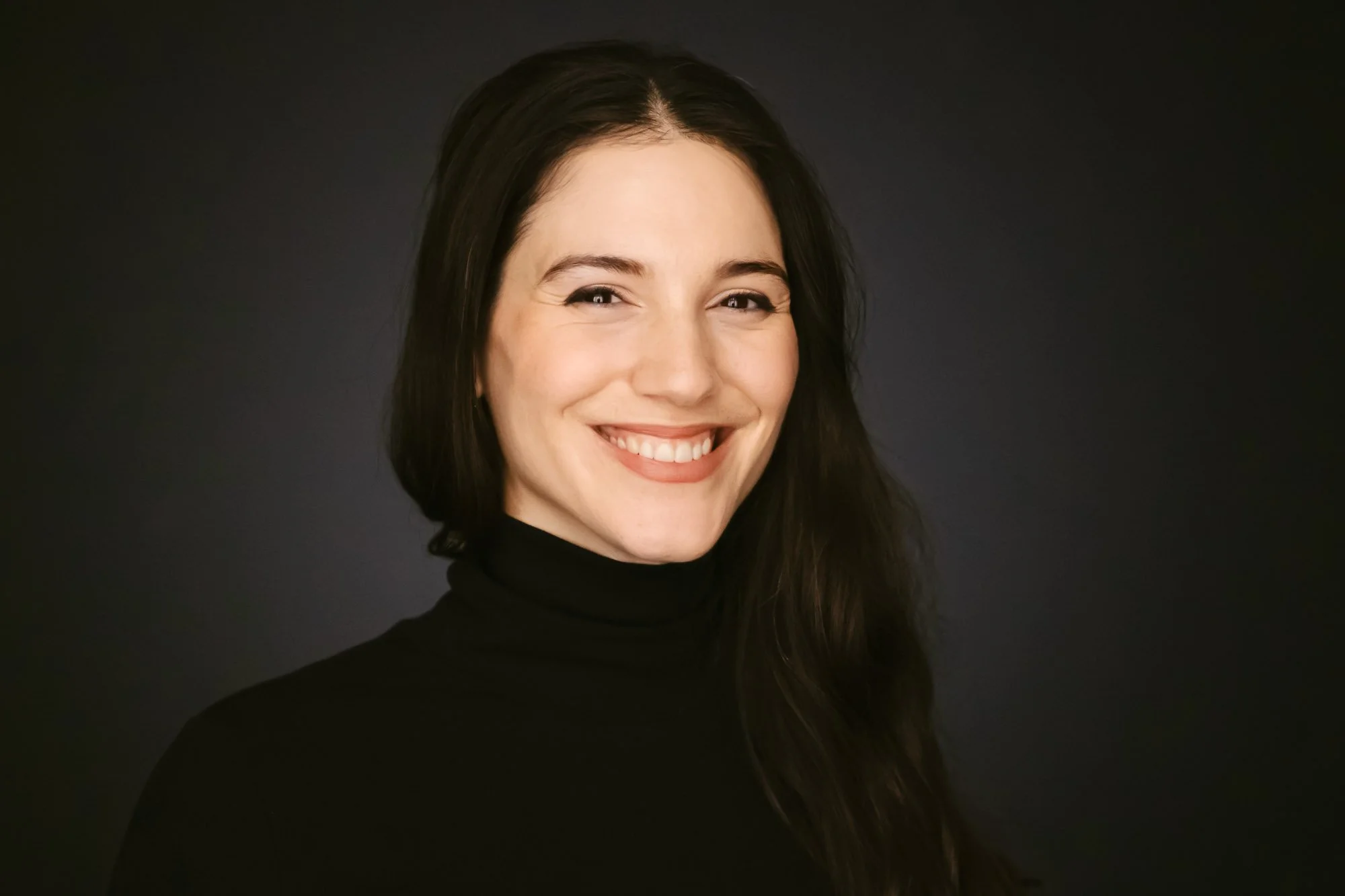 Danae Papadimitriou smiling, wearing a black turtleneck against a dark background.