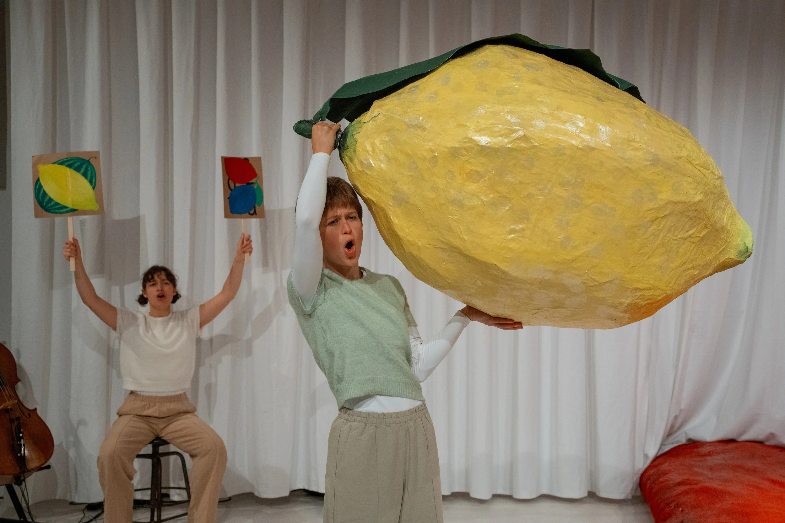 Two performers on stage with a white curtain backdrop: one person holding a large yellow prop shaped like a lemon, while another holds a small sign with a painted lemon design. The person with the lemon prop appears to be singing or speaking passiona