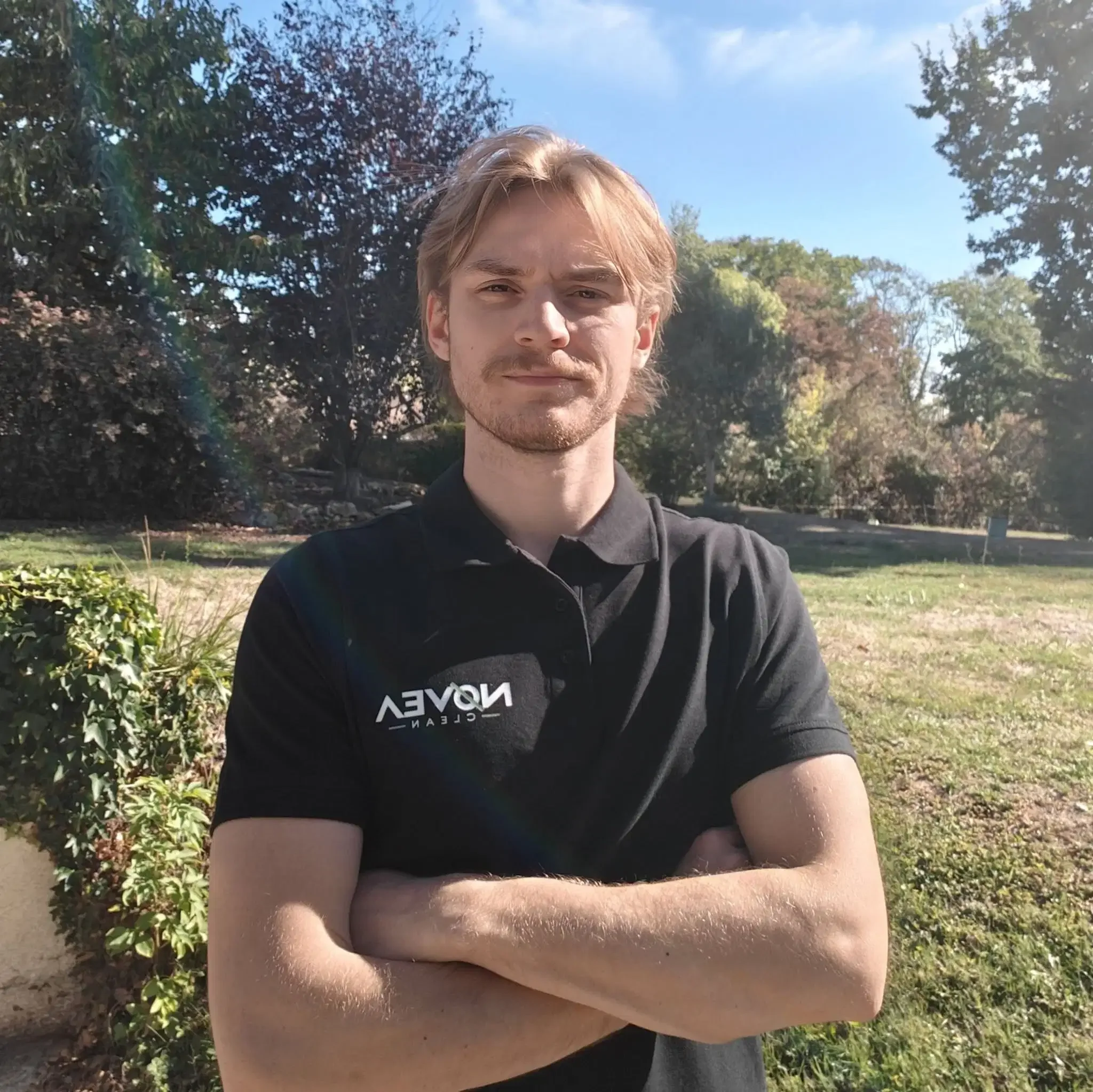 Lucas, fondateur de NOVEA CLEAN, jeune homme avec cheveux blonds et barbe légère, portant un t-shirt noi au logo de l'entreprise, prenant un selfie à l'extérieur sous un ciel bleu avec des arbres en arrière-plan.