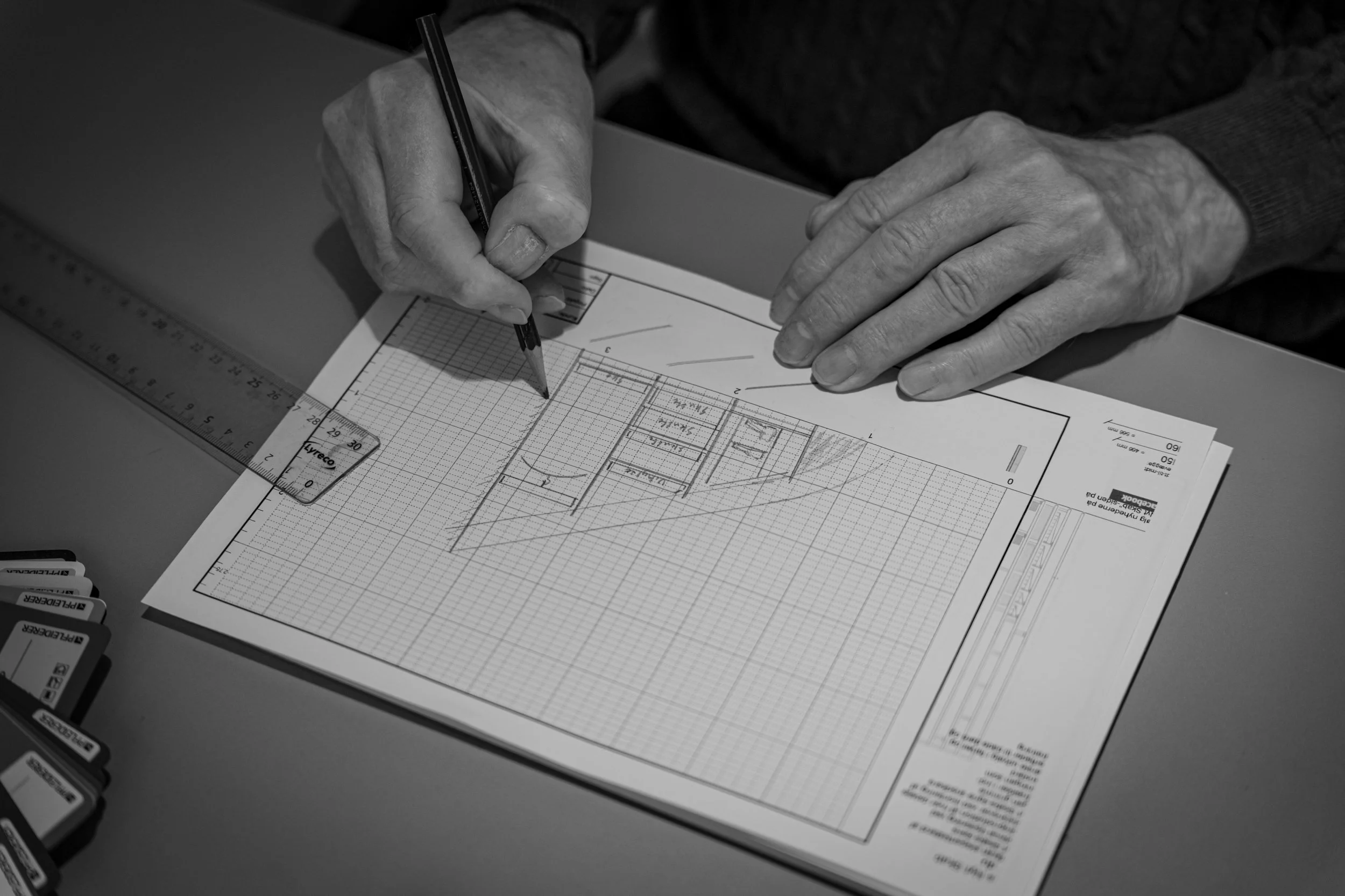 Person drawing architectural plans on graph paper with a pencil, ruler, and scale on a desk.