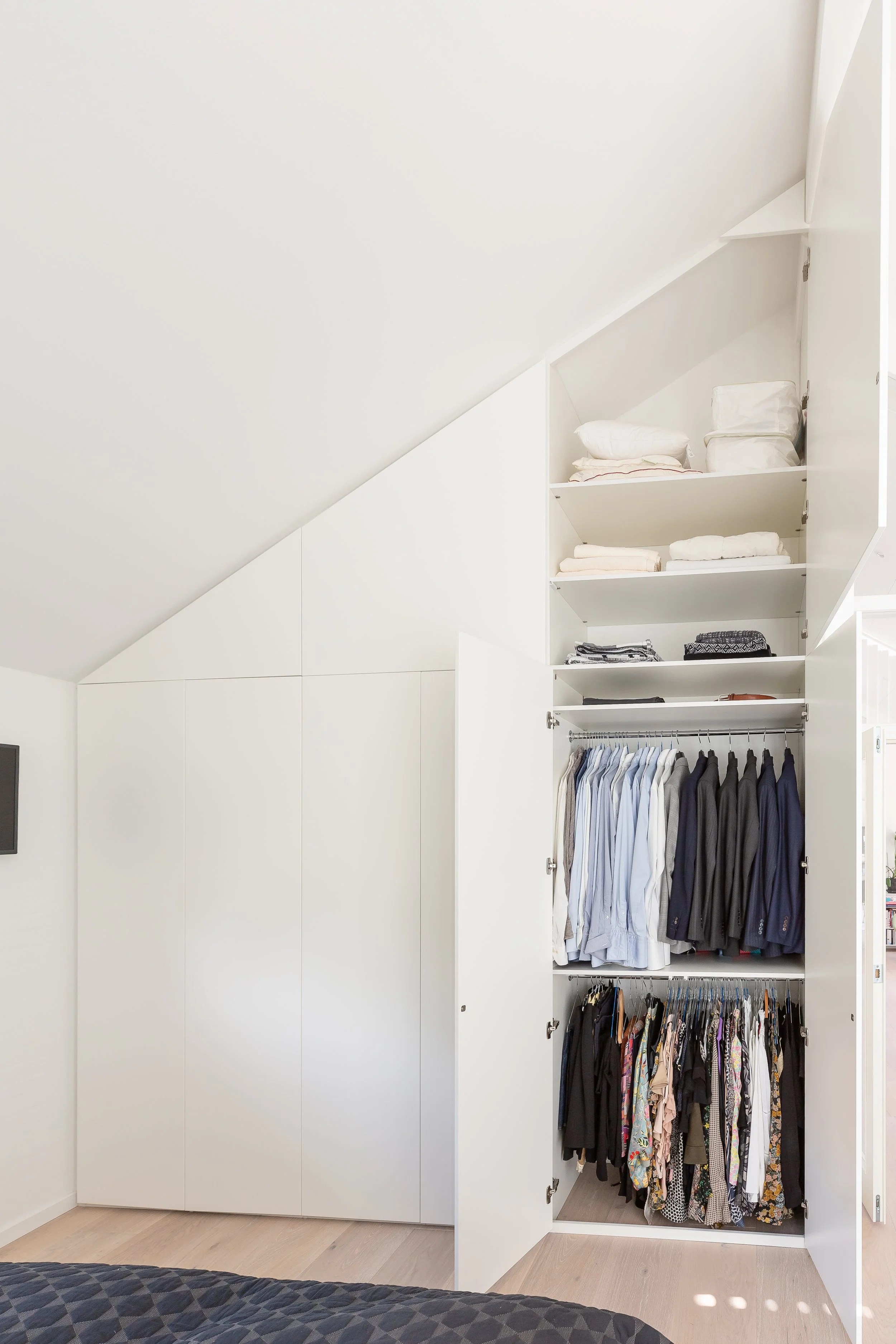 Open white closet with storage on an angled wall, containing folded clothes on shelves and hanging jackets and shirts.