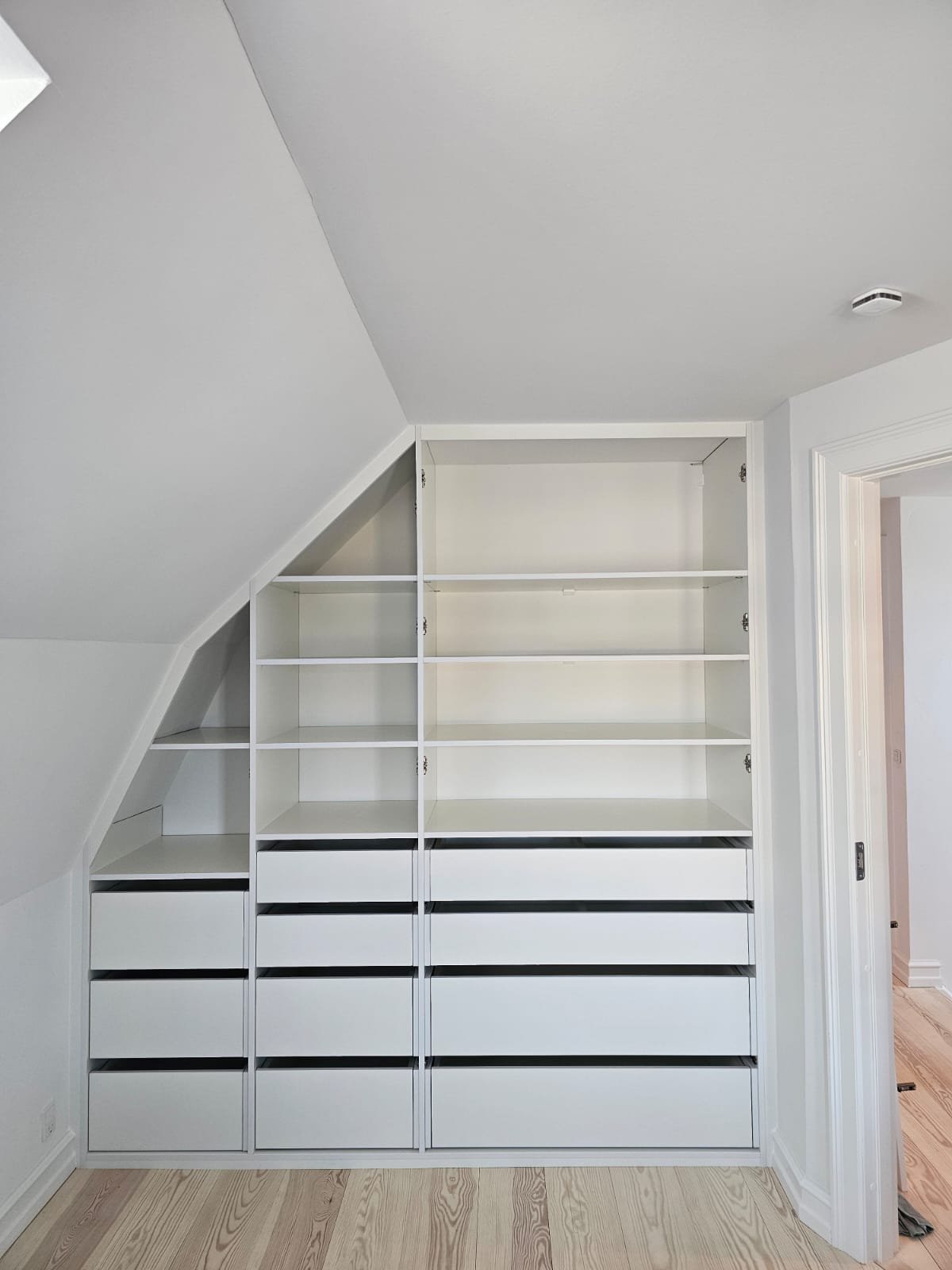 Empty white built-in shelving and drawers in a room with sloped ceiling.
