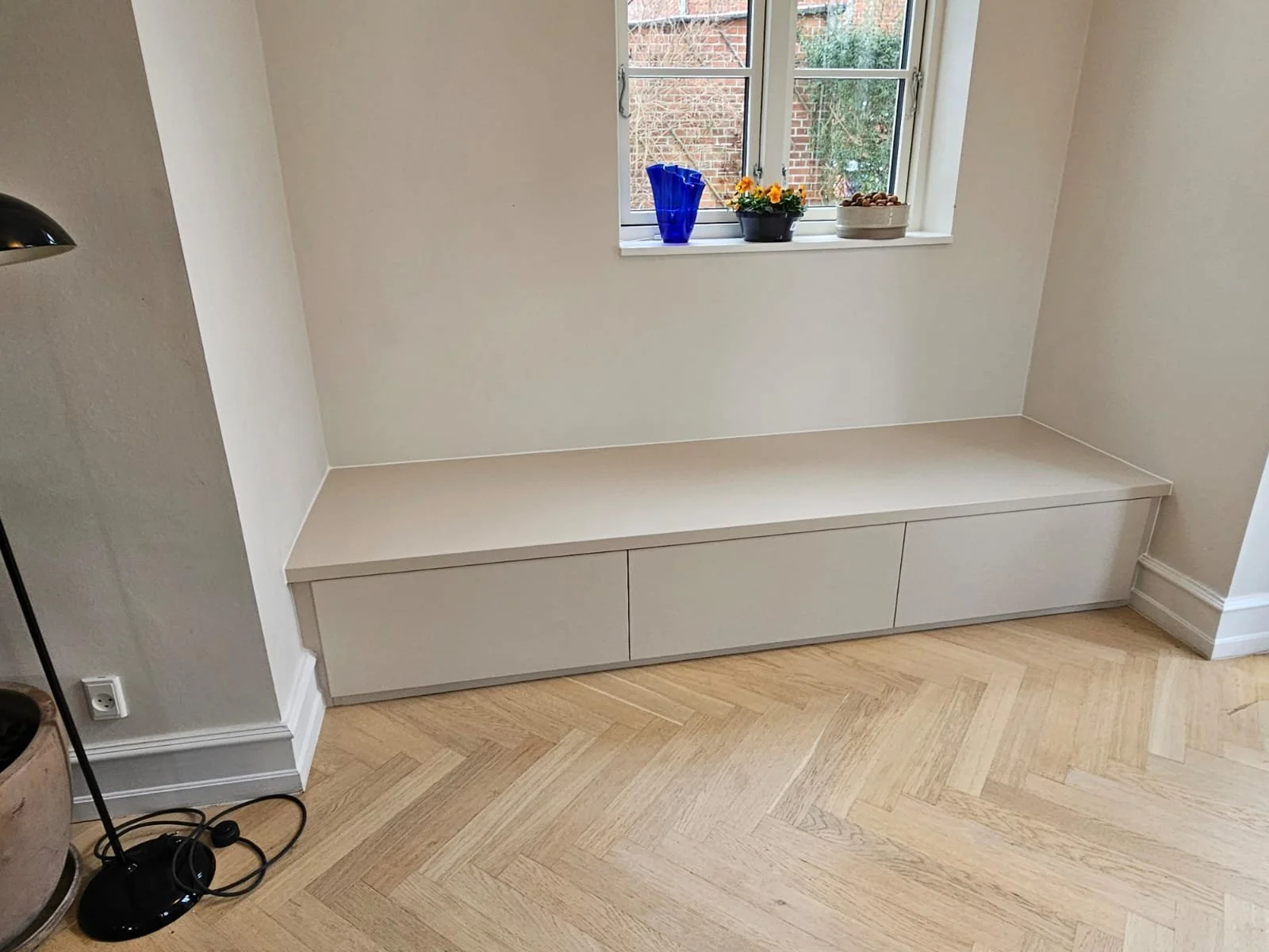 Built-in white storage bench beneath a window on a beige wall in a room with light hardwood flooring.