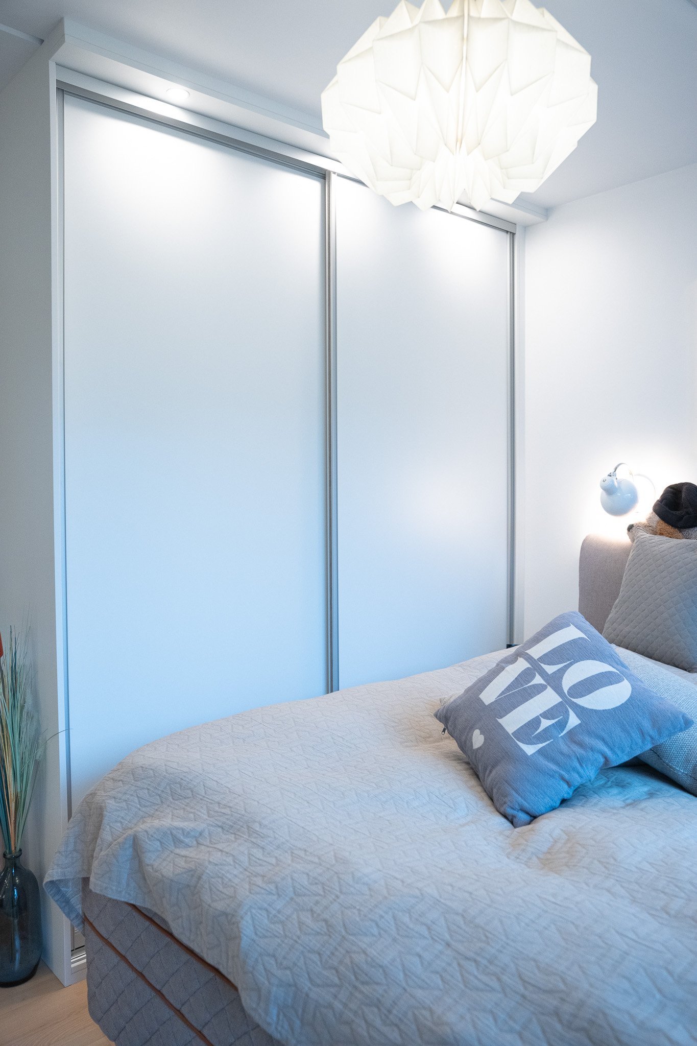 A bedroom with closed sliding closet doors, a bed with beige quilted bedding, grey pillow with 'LOVE' written on it, a bedside lamp, and a modern ceiling light fixture.