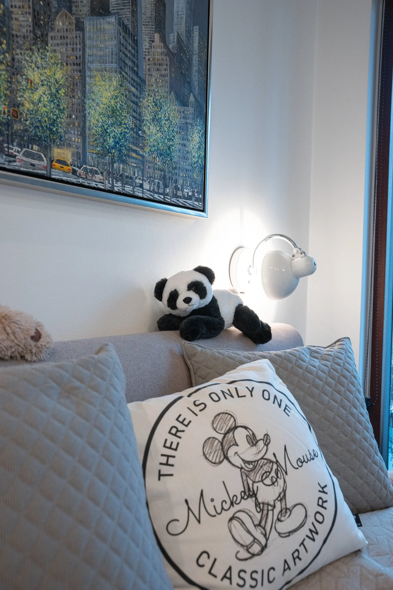 A cozy interior with a beige sofa adorned with patterned and plain cushions, a plush panda toy on the backrest, a painting of the city street scene hangs above, and a white wall-mounted lamp illuminates the space.