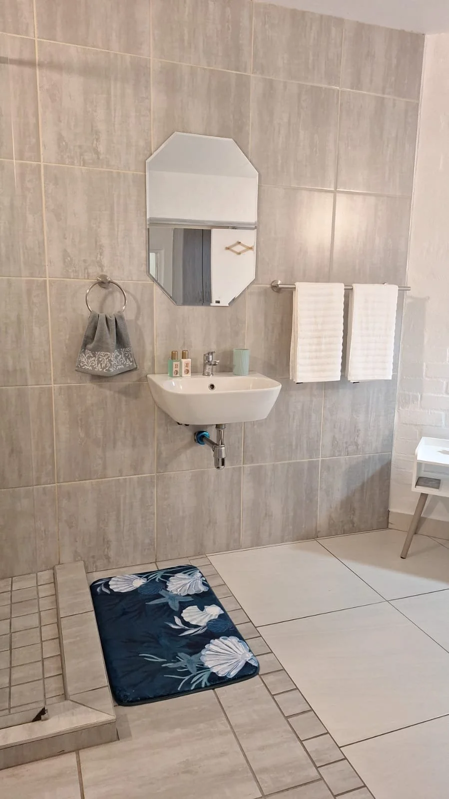 A bathroom with a small wall-mounted sink, a mirror above it, a towel ring with a gray hand towel on the left, two white towels on a towel bar to the right, a small white side table, a navy blue floral bath mat on tiled floor, and tiled walls.