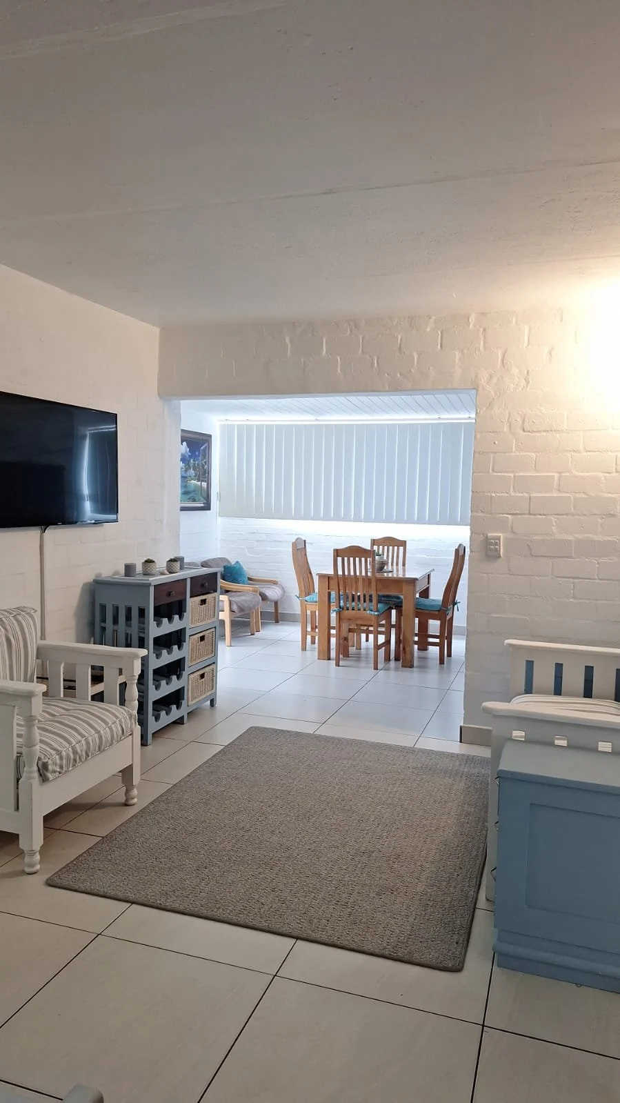 Living room and dining area with white brick walls, tiled floor, a wall-mounted TV, blue and white furniture, a beige rug, and a dining table with six chairs near a window with closed blinds.