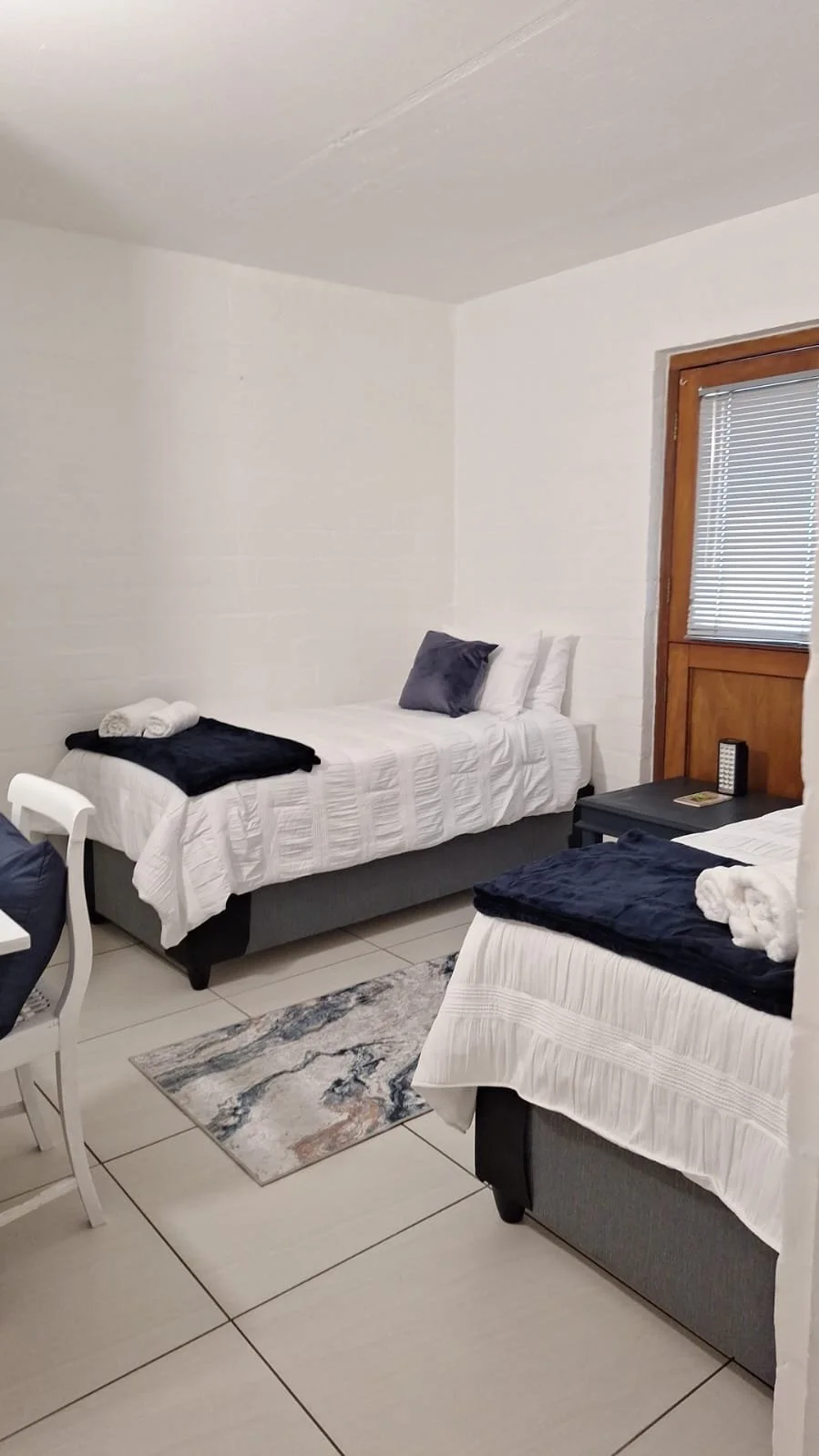A bedroom with two single beds, white bedding, gray pillows, a dark side table with a phone and remote, a window with blinds, a small colorful rug, and towel rolls on the beds.