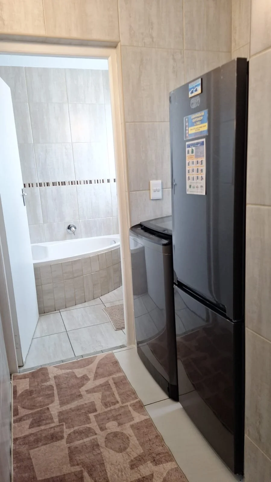 View of a laundry area with a black refrigerator, a small washing machine, beige tiled walls, and a doorway leading to a bathroom with a bathtub.