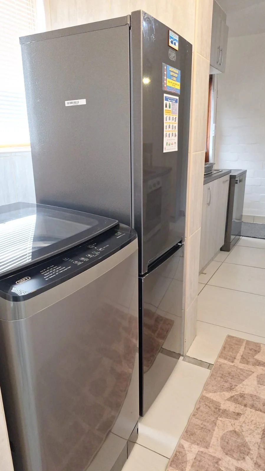 A laundry room with a stainless steel washing machine and a gray refrigerator side by side, with beige tiled flooring and a light-colored wall.