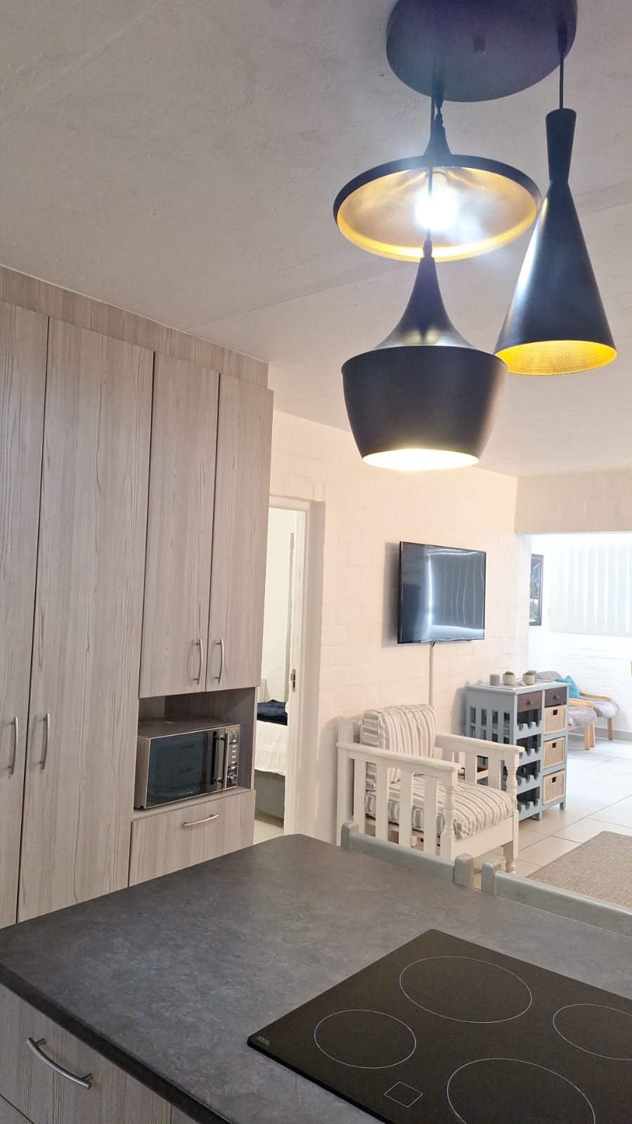 Modern kitchen with black pendant lights hanging from ceiling, wood cabinets, microwave built into cabinetry, and a dining area with TV on the wall and striped armchair in background.