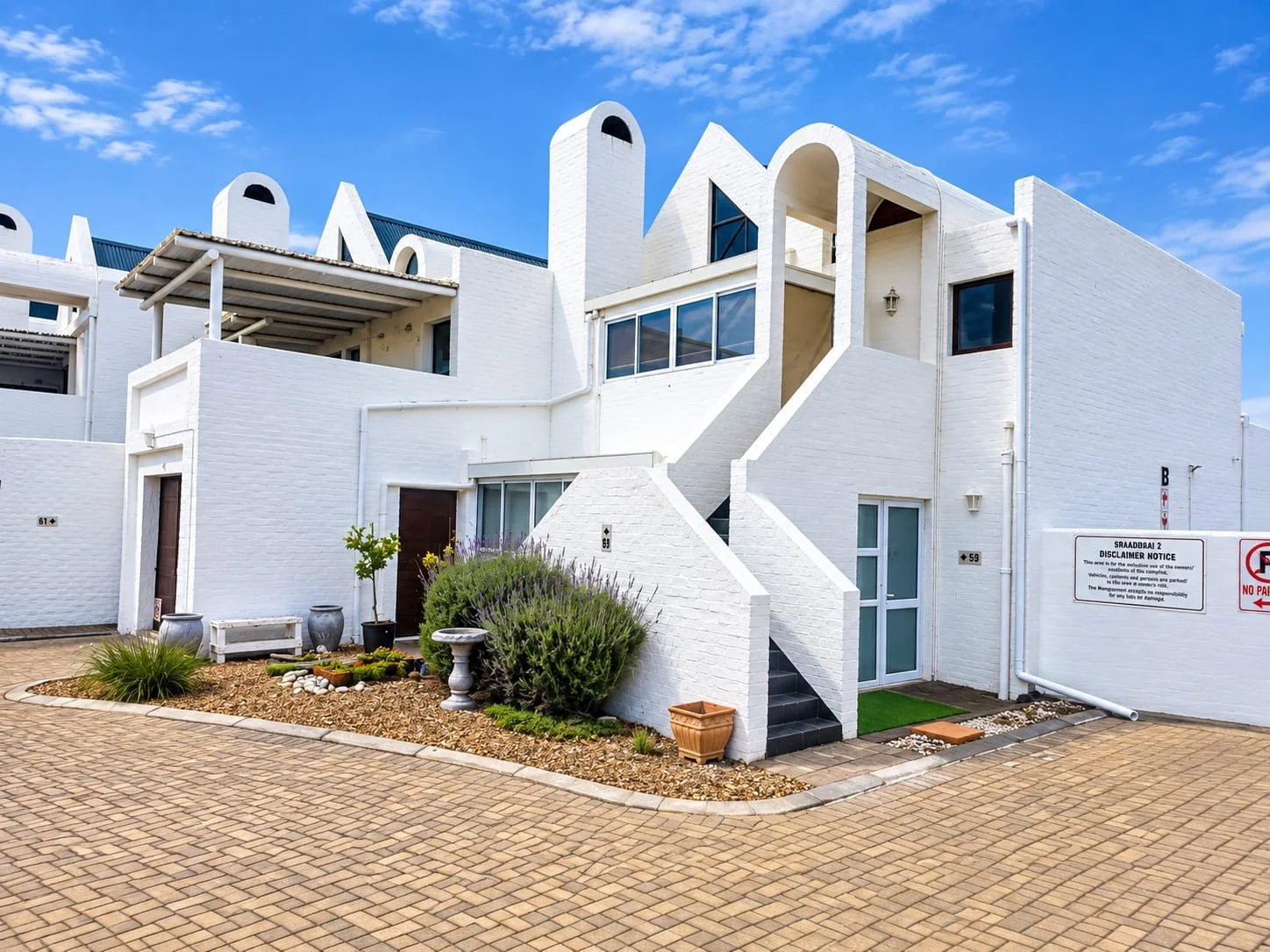 White modern multi-story residential building with curved architectural elements, small garden, potted plants, and a paved pathway, under a partly cloudy blue sky.