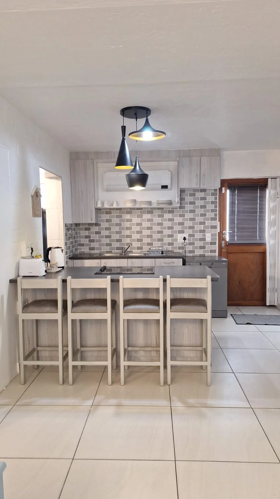 Modern kitchen with light-colored cabinets, gray countertops, a tiled backsplash, and a breakfast bar with four chairs. Pendant lights hang from the ceiling. There is a door with blinds on the right side.