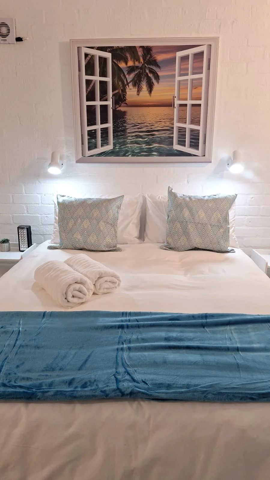A neatly made bed with two patterned pillows, white towels, and a blue blanket at the foot of the bed. Above the bed is a mural of a tropical sunset with palm trees and a water view, designed to look like a window.