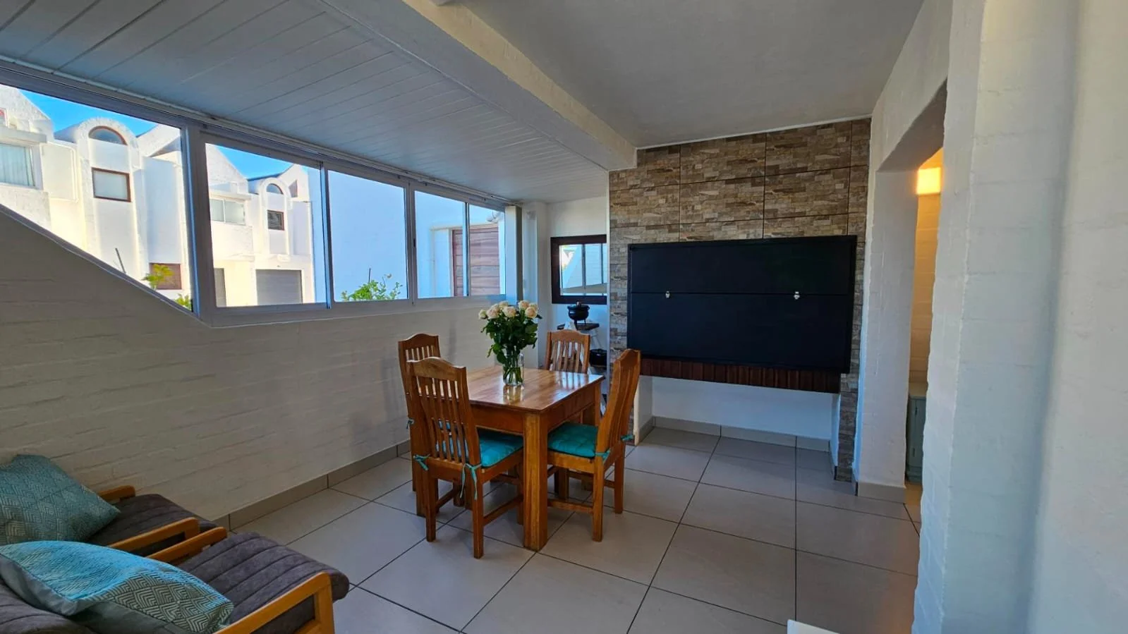 Living room with large window, wooden dining table with four chairs, a vase of flowers, wall-mounted television, brick accent wall, and white painted brick walls.