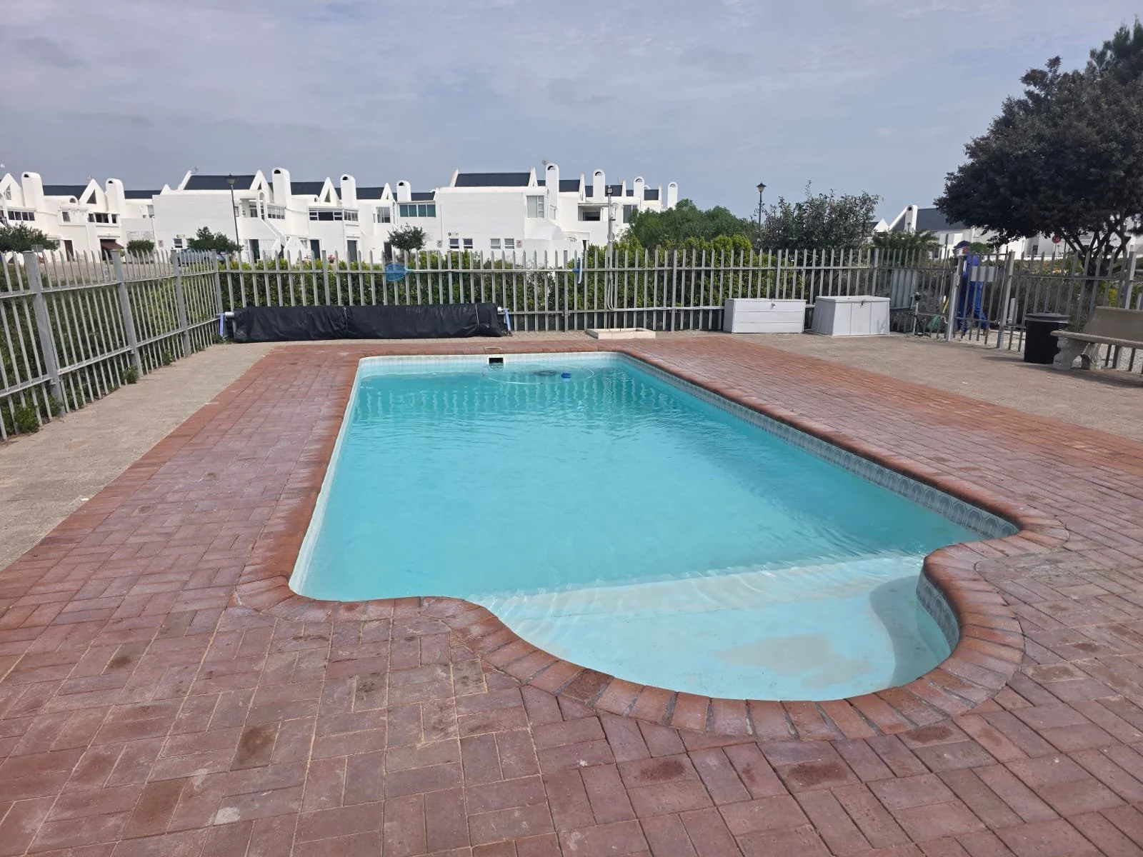 An outdoor swimming pool with clear blue water, surrounded by a brick patio and a white fence. In the background, there are white residential buildings, a large tree on the right, and some outdoor furniture.