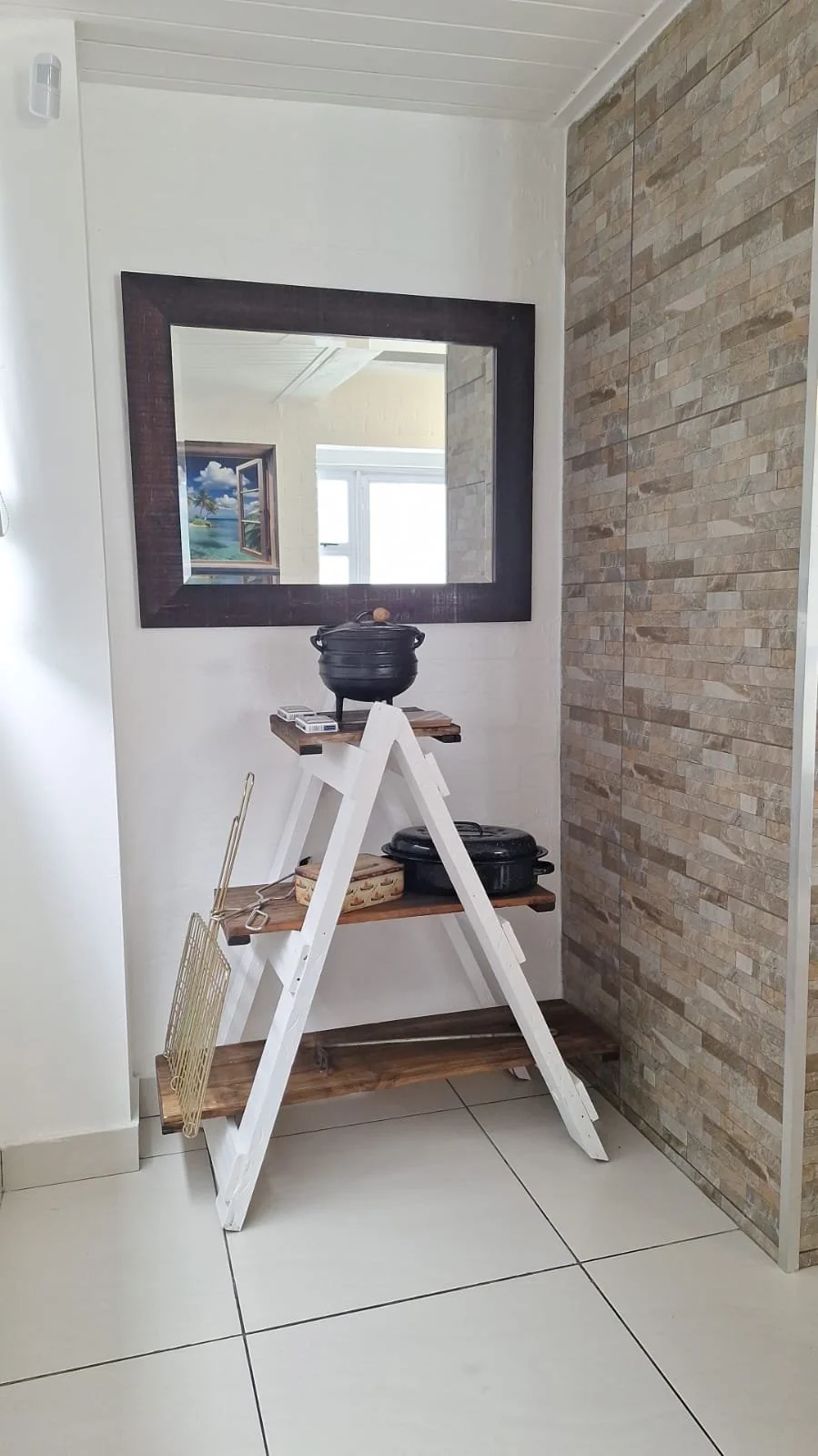 A white A-frame shelf with wooden shelves holding black cast iron pots and a small woven basket, placed in a room with tiled floor, a wall with stone tiles, and a large mirror reflecting a window and scenery outside.
