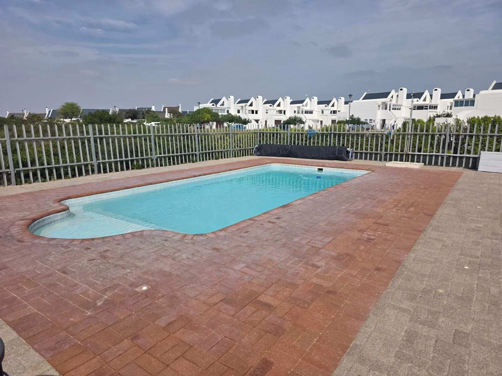 Empty swimming pool with a brick deck, surrounded by a metal fence, and white buildings in the background under a partly cloudy sky.
