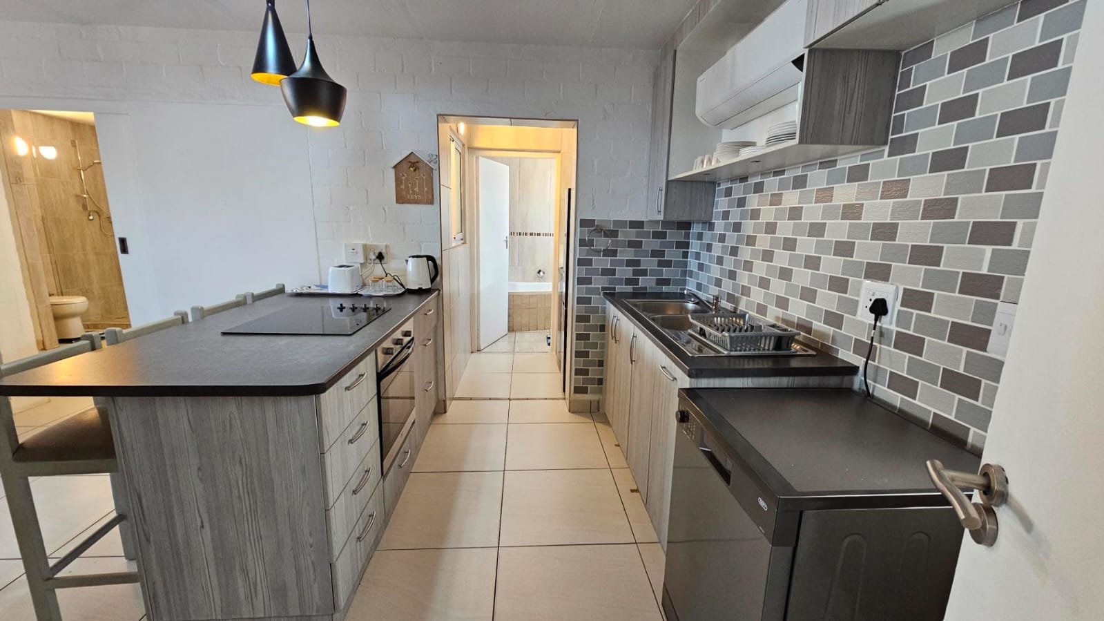 A modern kitchen with gray cabinetry, a gray-tiled backsplash, a double sink, and appliances including a dishwasher, stovetop, oven, and kettle. There is a small table with chairs and an open door leading to a bathroom with a bathtub visible.